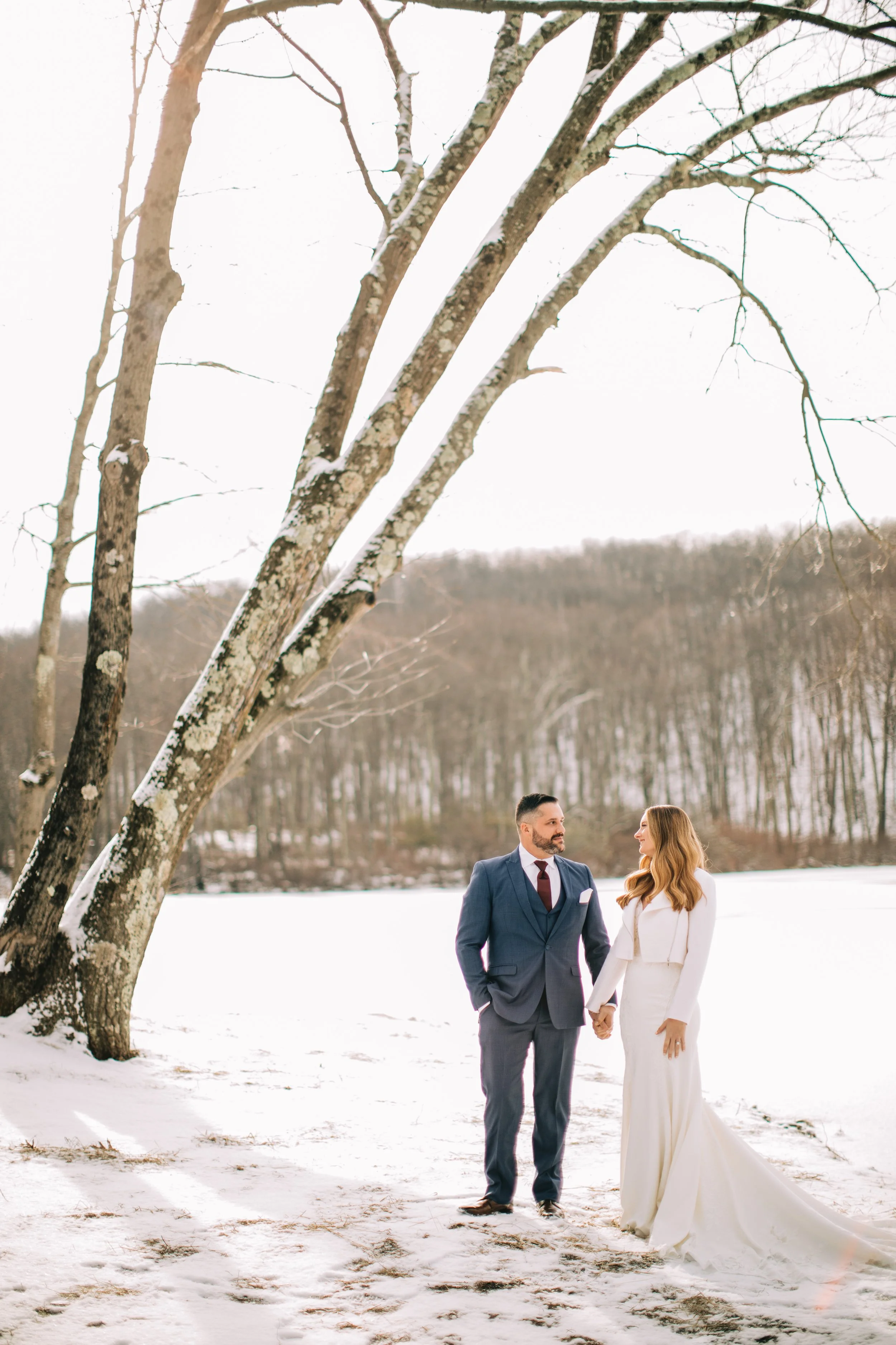 A man and woman in wedding attire holding hands in a snowy landscape with a large leafless tree and a forested hill in the background.