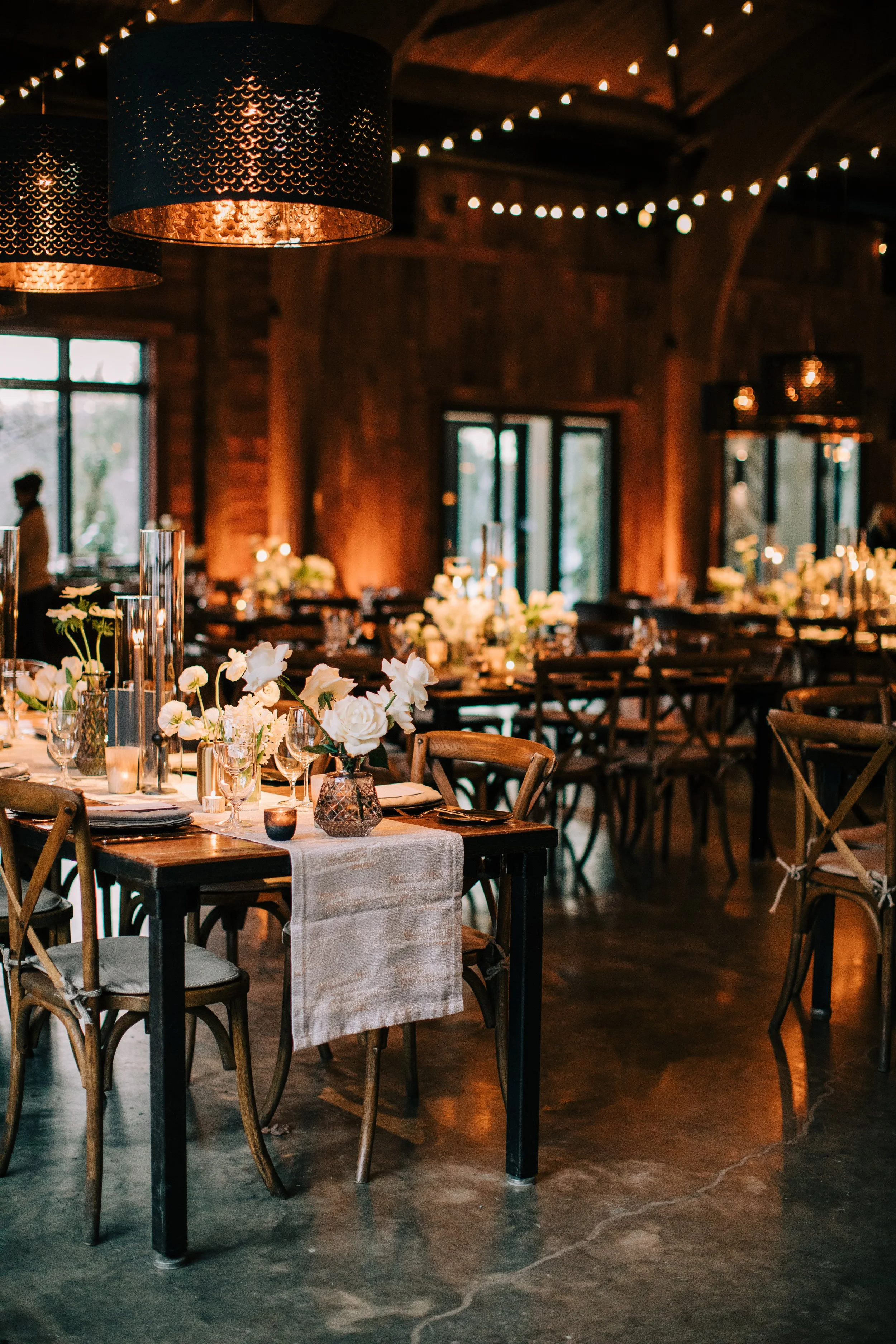 Elegant indoor wedding reception with round tables decorated with white flowers, candles, and glassware, featuring warm lighting and a rustic wooden interior.
