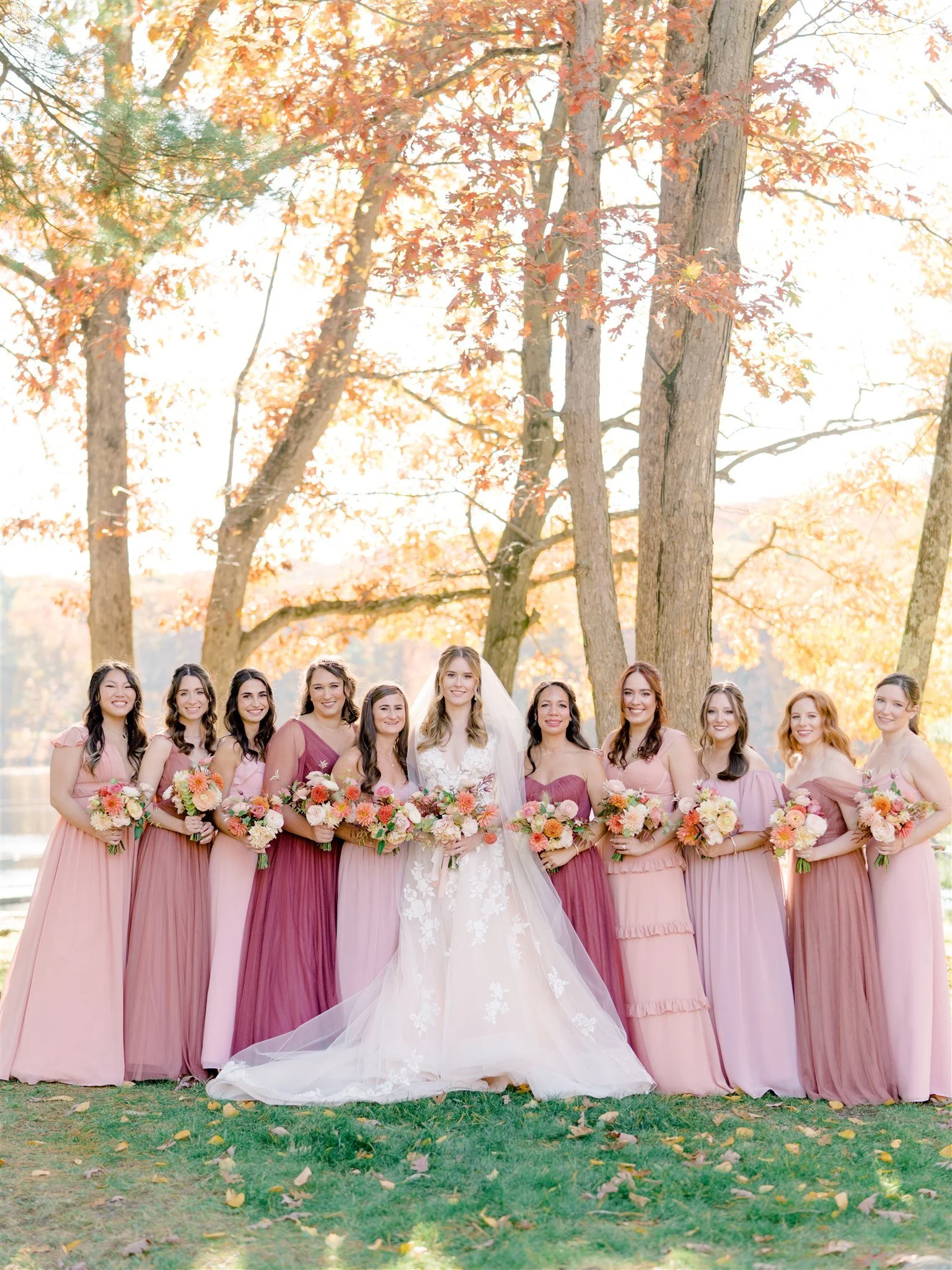 A bride and ten bridesmaids standing outdoors in a park with fall foliage, holding bouquets of pink, peach, and white flowers.