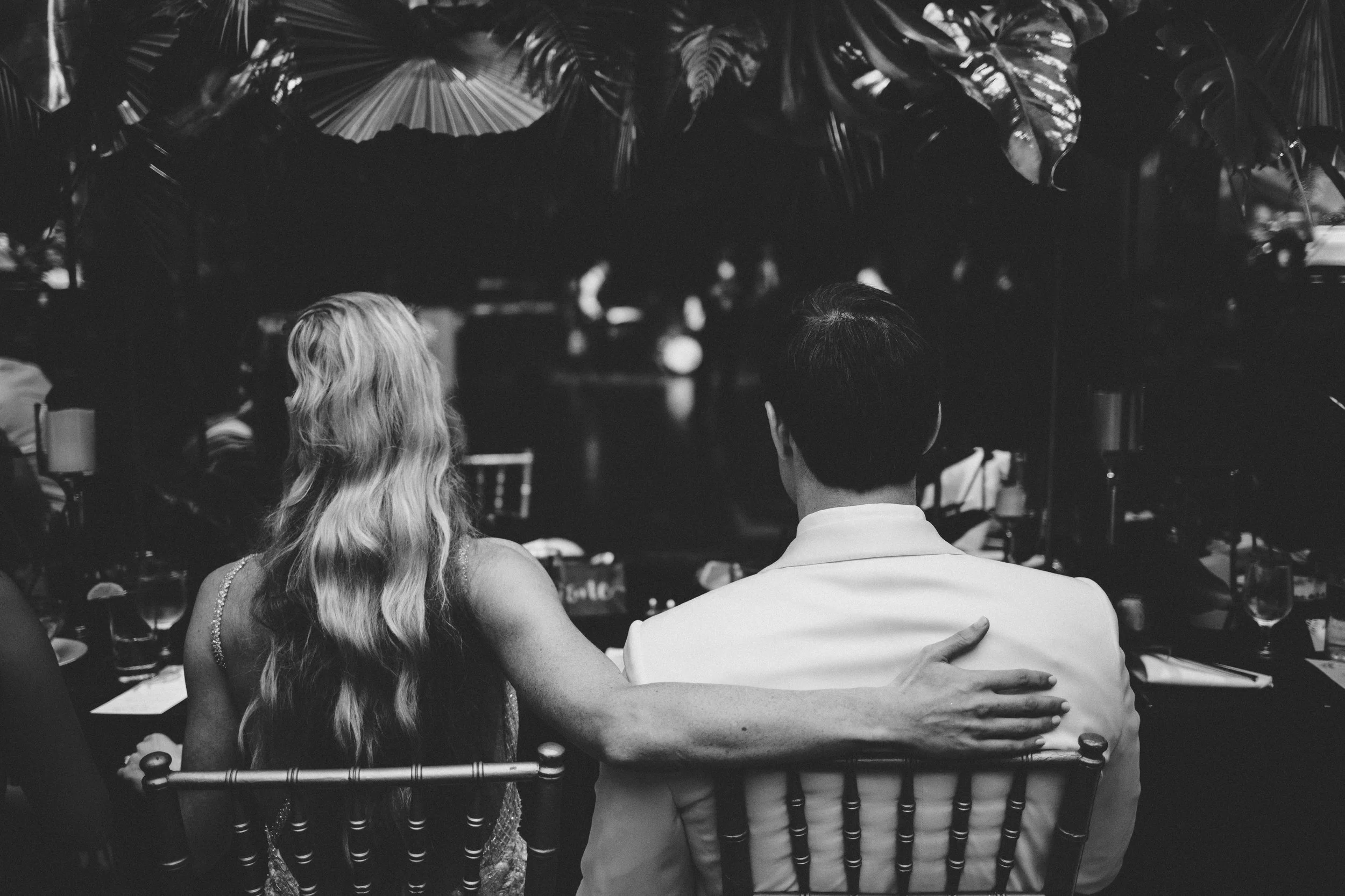 A black and white photo of a man and woman sitting together at a formal event, with the woman's arm around the man's shoulder, viewed from behind.
