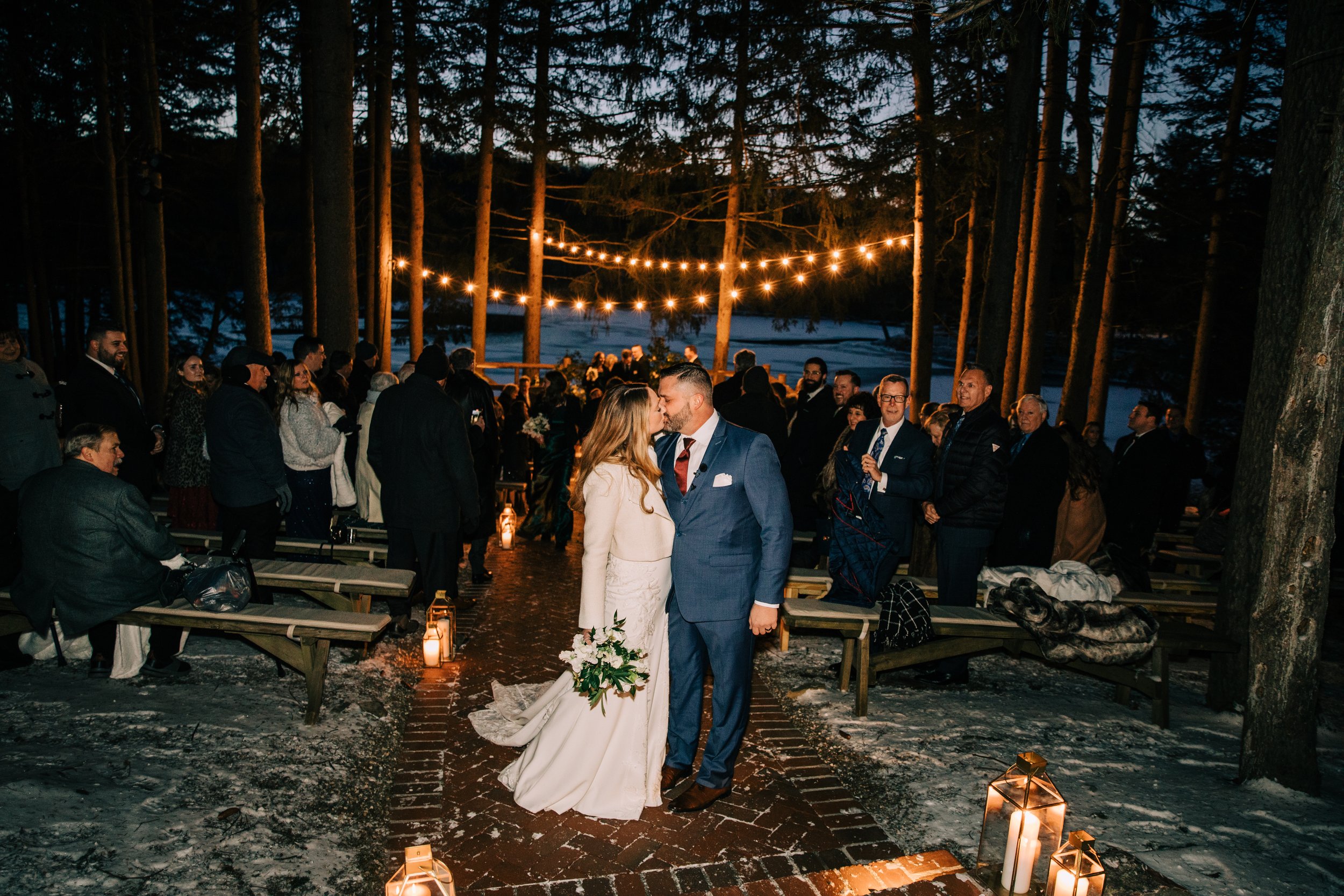 A wedding couple shares a kiss during their ceremony outdoors at night, surrounded by guests in a forest setting with string lights overhead and lanterns on the ground lining a brick aisle.