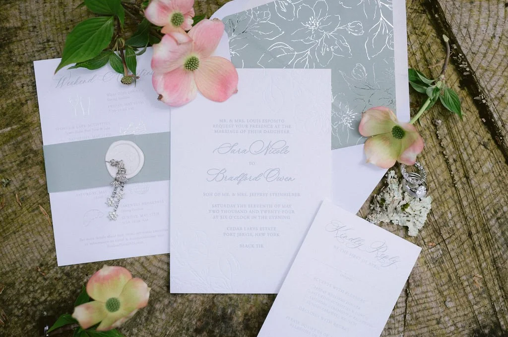 Wedding invitation suite with floral accents and jewelry, laid on a rustic wooden surface.