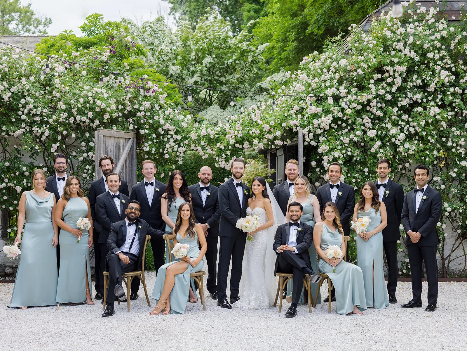 A wedding party with bridesmaids and groomsmen posing outdoors under flowering bushes, with some seated and others standing, for a group photo.