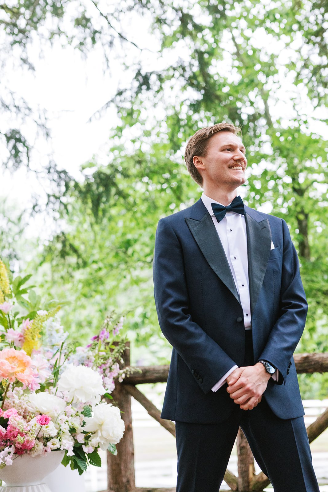 Man in a tuxedo standing outdoors near colorful flowers, smiling and looking to the side.