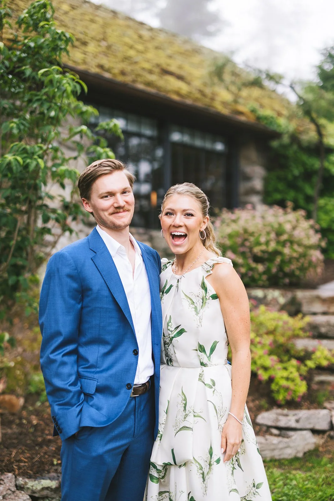 A smiling couple standing outdoors in front of a house with a mossy roof and lush greenery. The man is dressed in a blue suit, and the woman is wearing a white dress with green leaf patterns.