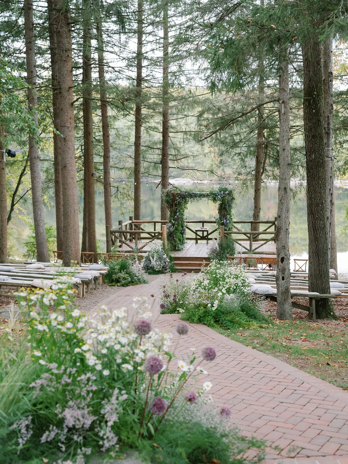 A peaceful outdoor wedding setup in a wooded area with a decorated arch at a lakeside, surrounded by trees, flowers, and benches.