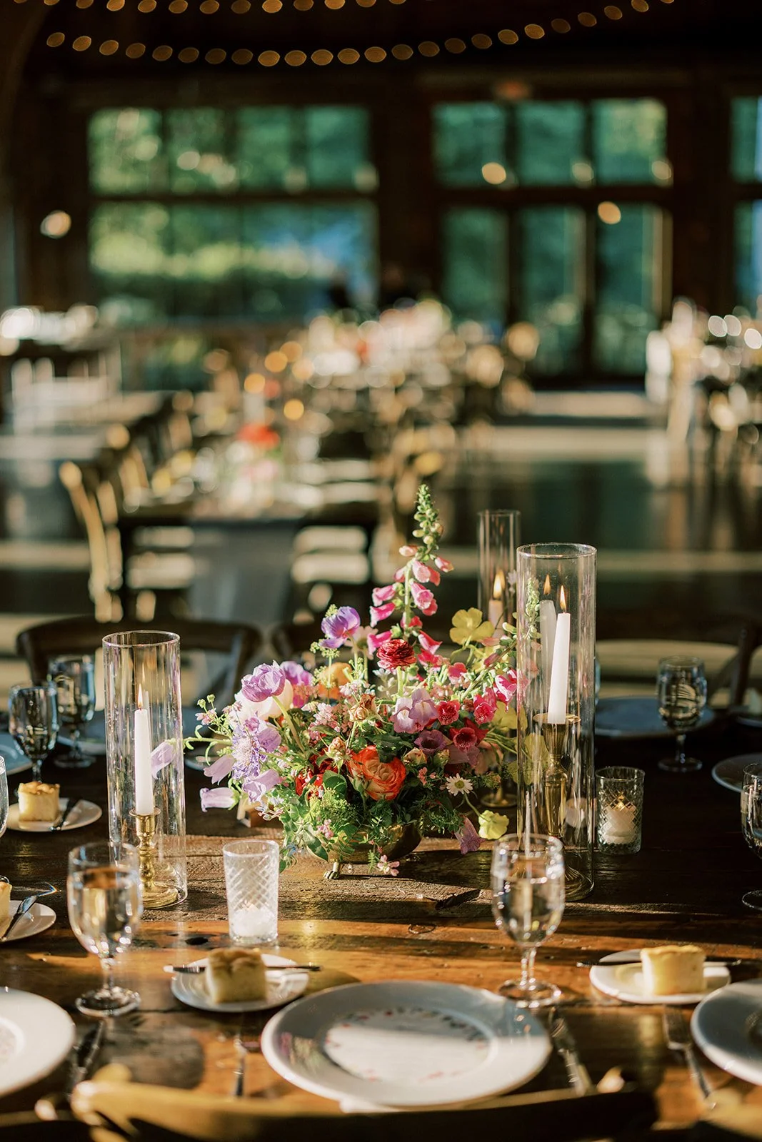 Decorated dining table with a colorful flower arrangement, candles in glass holders, and place settings, set in a rustic, sunlit hall with large windows.