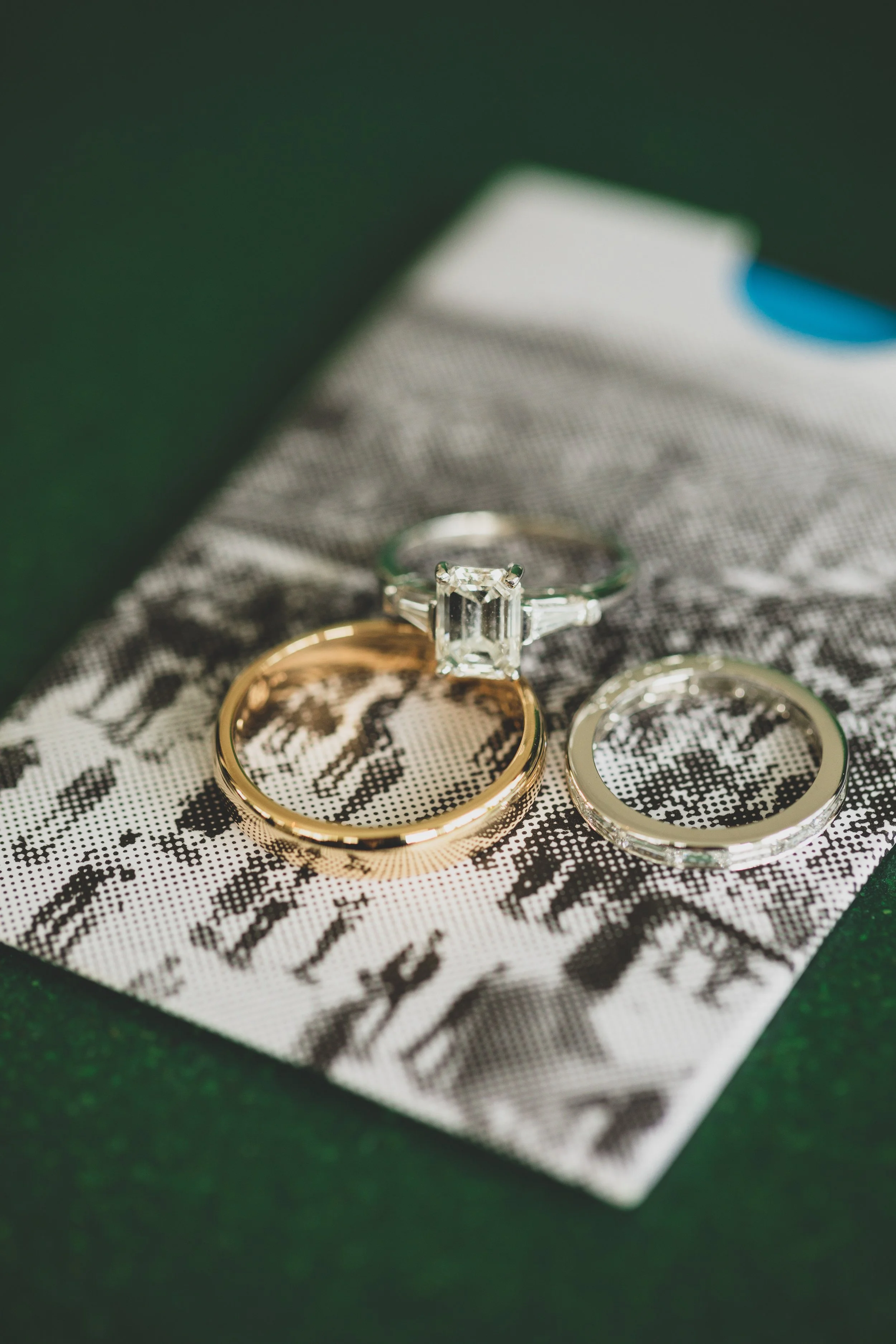 Two rings, one gold and one silver, with the gold ring featuring a large emerald-cut diamond, placed on a patterned fabric with black and white abstract design.