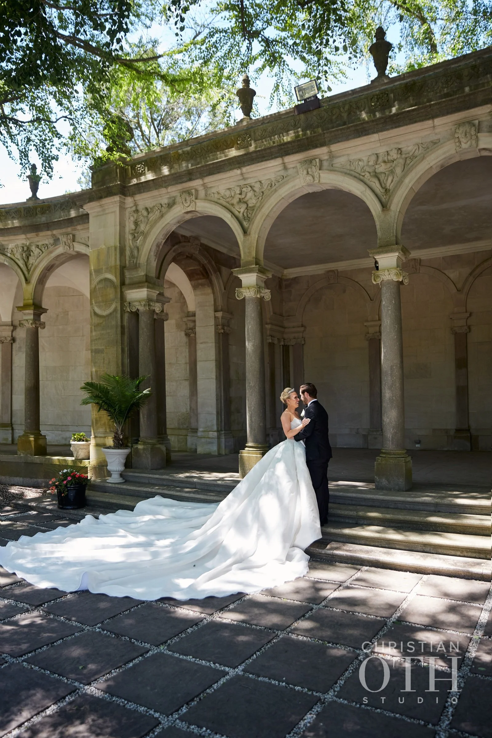 Bride and groom dancing outdoors under shaded arches, with the bride in a long white gown with a train, and the groom in a black tuxedo, in a courtyard with stone steps and potted plants.