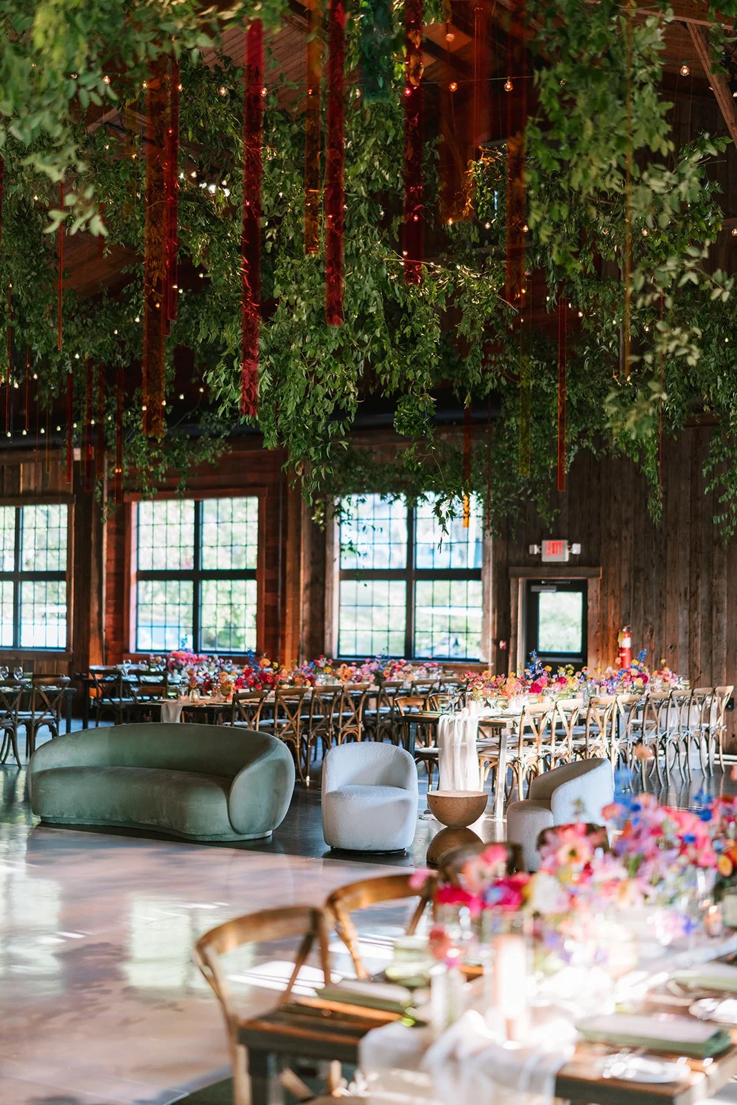 Intimate indoor event space decorated with hanging greenery and ribbons, long dining tables with colorful floral centerpieces, and modern seating including a green velvet sofa and white chairs.