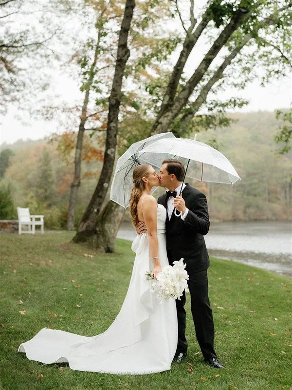 A newlywed couple sharing a kiss under a transparent umbrella outdoors near a lake, with trees in the background and a white bench nearby.