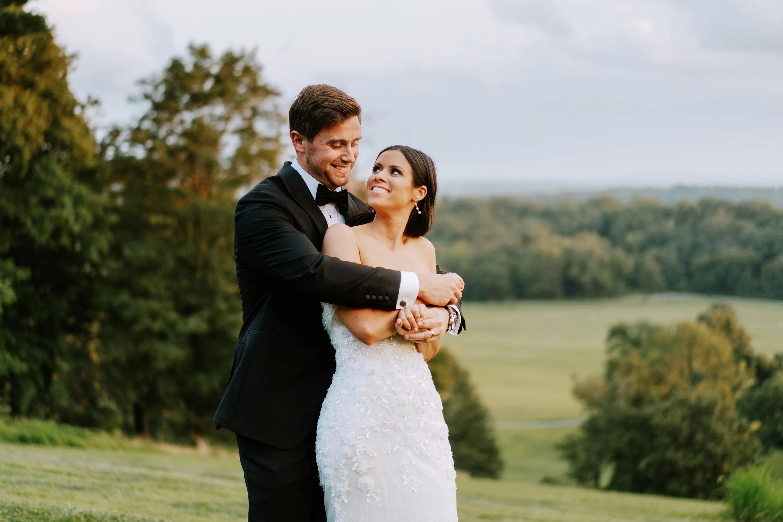 A newlywed couple in wedding attire standing outdoors on a grassy hill, with trees and a scenic landscape in the background. The groom in a black tuxedo hugs the bride in a white wedding gown, both smiling lovingly.