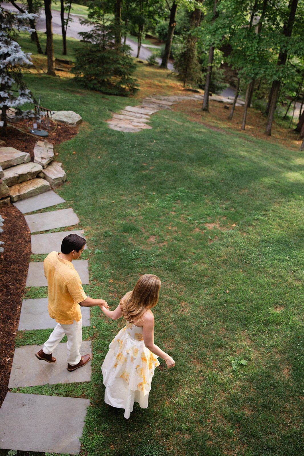 A young man and woman are holding hands while walking on a stone pathway in a lush green backyard with trees and grass.