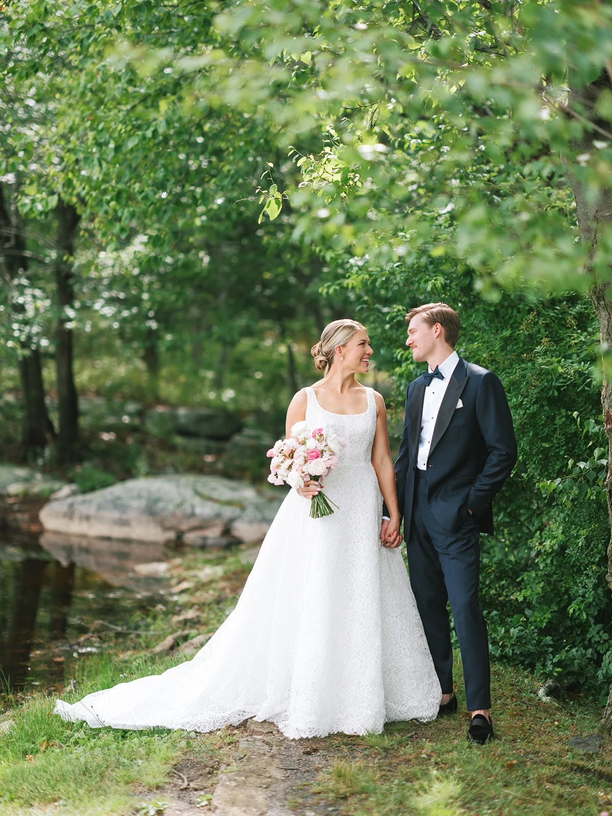 A newlywed couple stands together in a lush green outdoor setting, holding hands and gazing at each other, with the bride holding a bouquet of pink and white flowers.