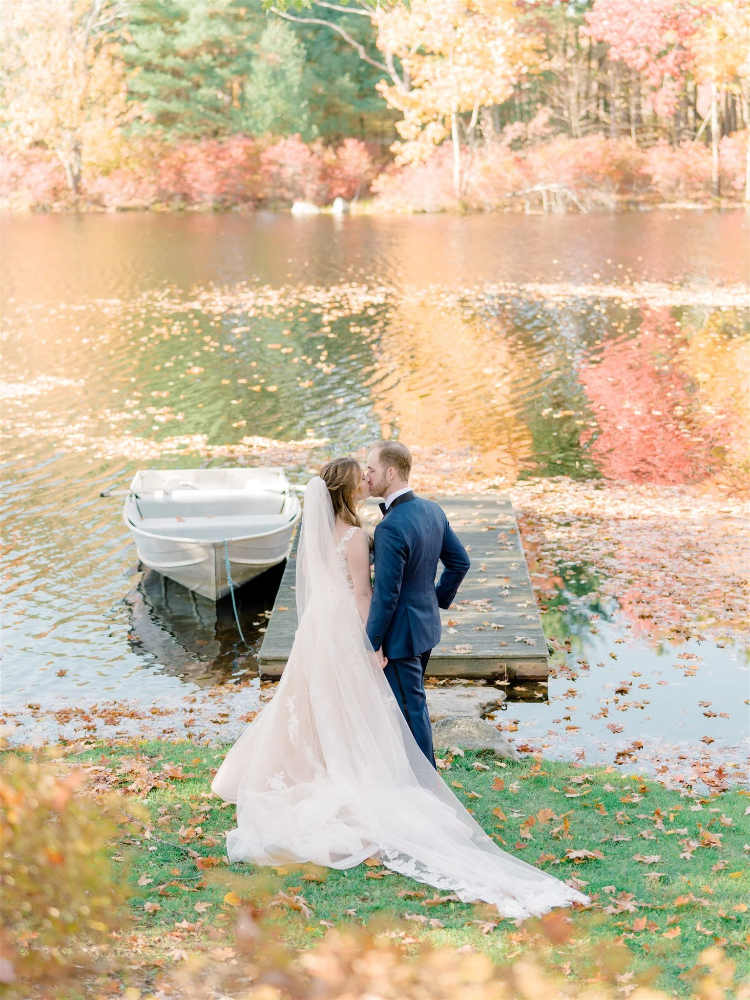 A bride and groom share a kiss on a dock beside a lake surrounded by fall trees and leaves.