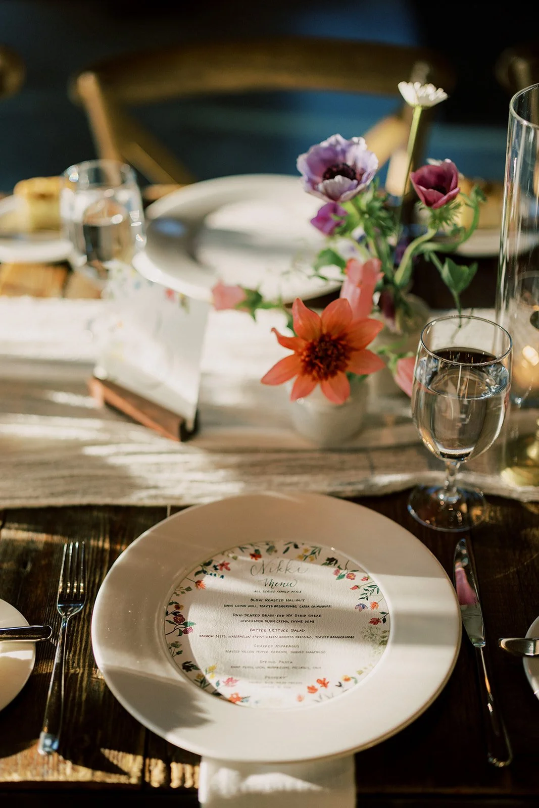 A beautifully set dining table with a floral centerpiece, a dinner menu, glass of water, plates, utensils, and a rustic wooden table.