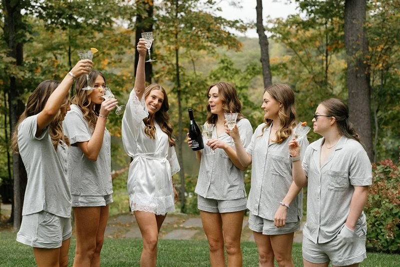 Six women wearing matching pajamas celebrating outdoors with glasses of champagne in a wooded area.