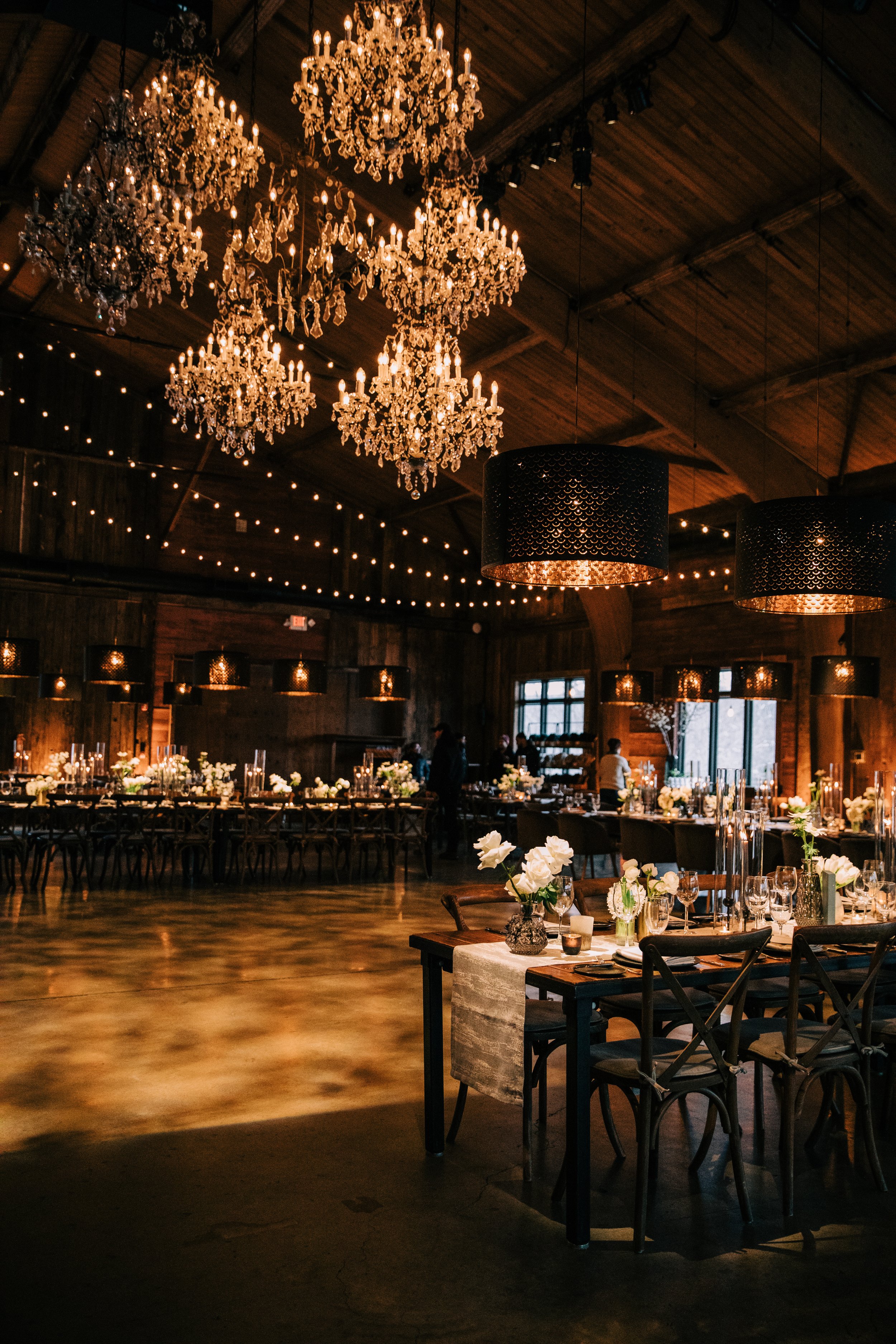 Elegant event hall with multiple chandeliers hanging from a wooden ceiling, decorated with string lights. Long tables are set with flower arrangements, candles, and glassware, suggesting a formal celebration or wedding reception.