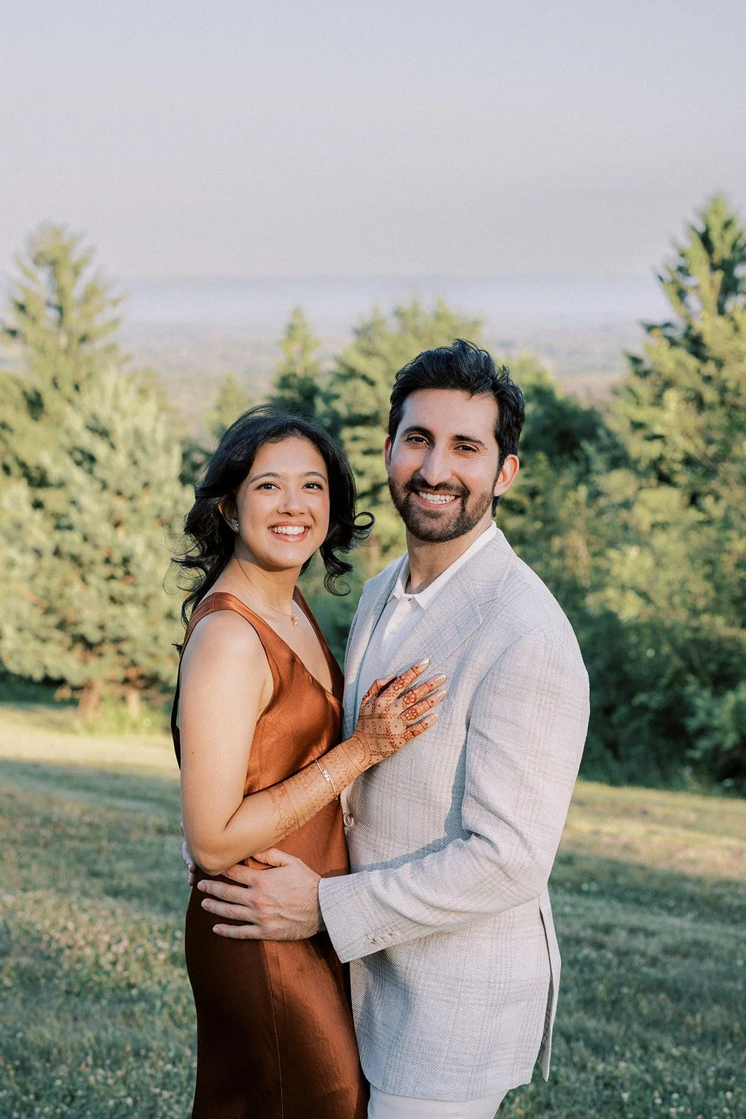 A smiling couple dressed in formal attire pose outdoors in a park with trees and a distant view of a landscape.