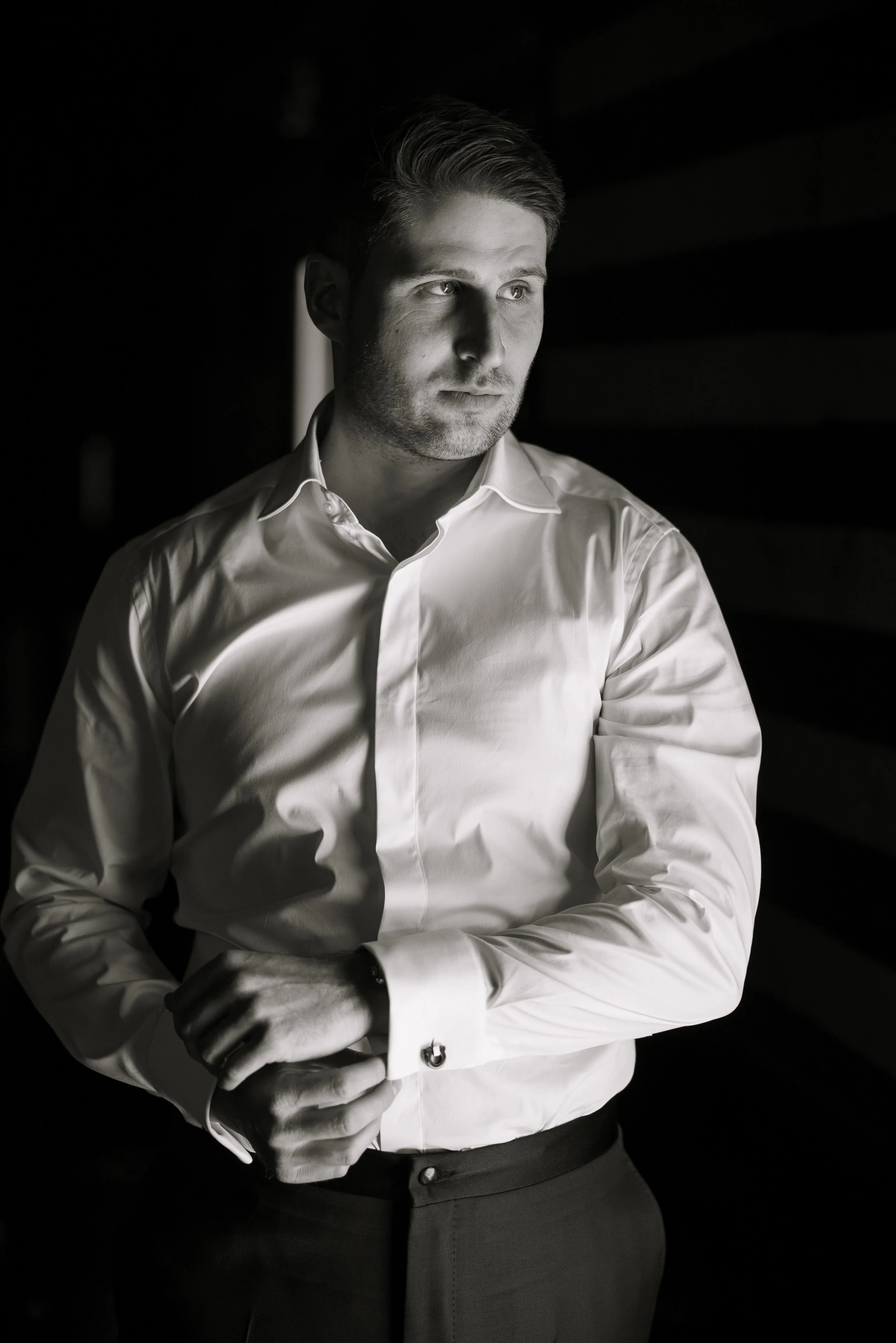 Black and white portrait of a young man in a white dress shirt adjusting his cuff, looking thoughtfully to the left, with shadows on his face.
