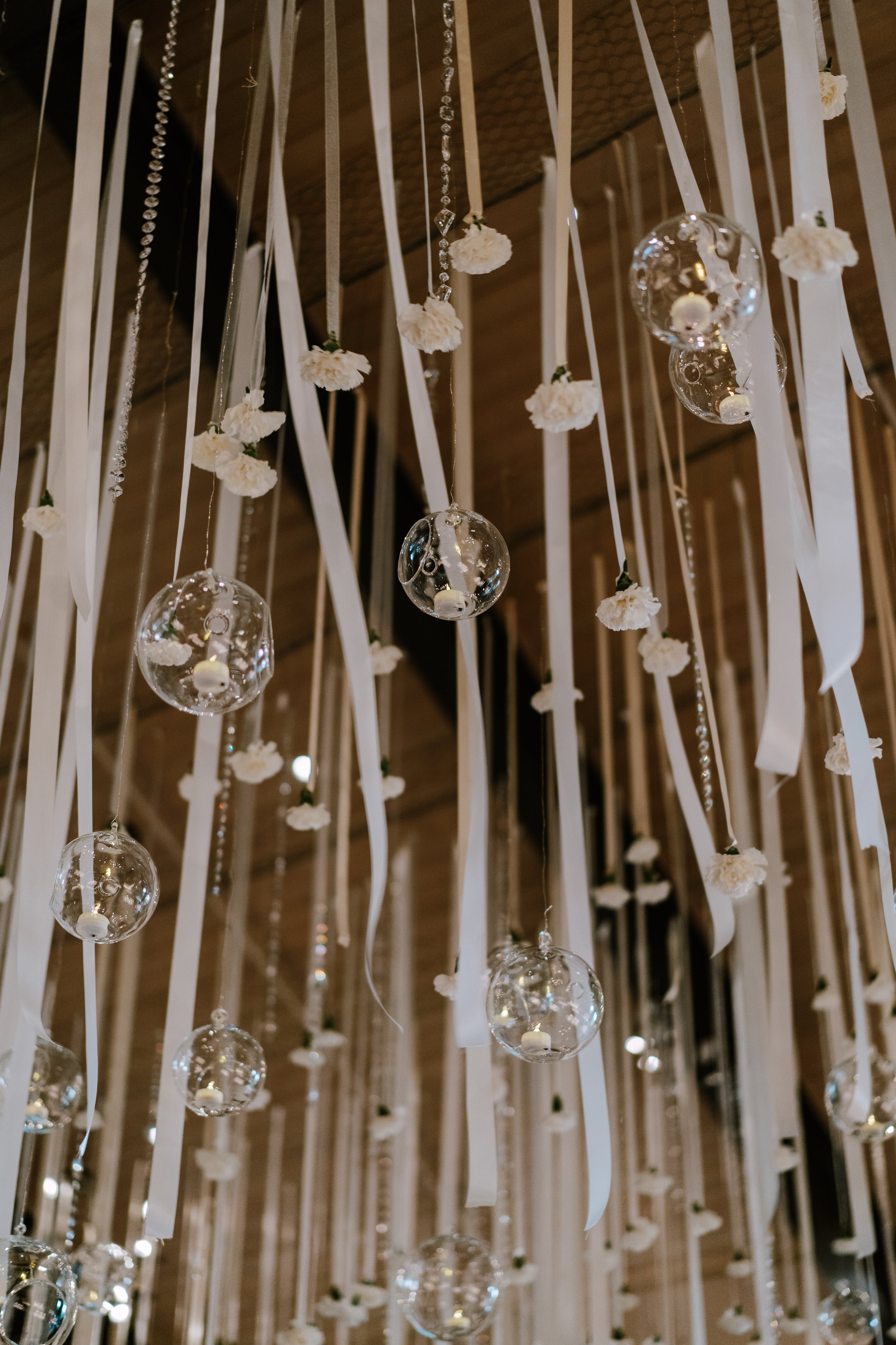 Decorative hanging display with white ribbons, small white flowers, and glass orbs containing candles, arranged in a vertical pattern.