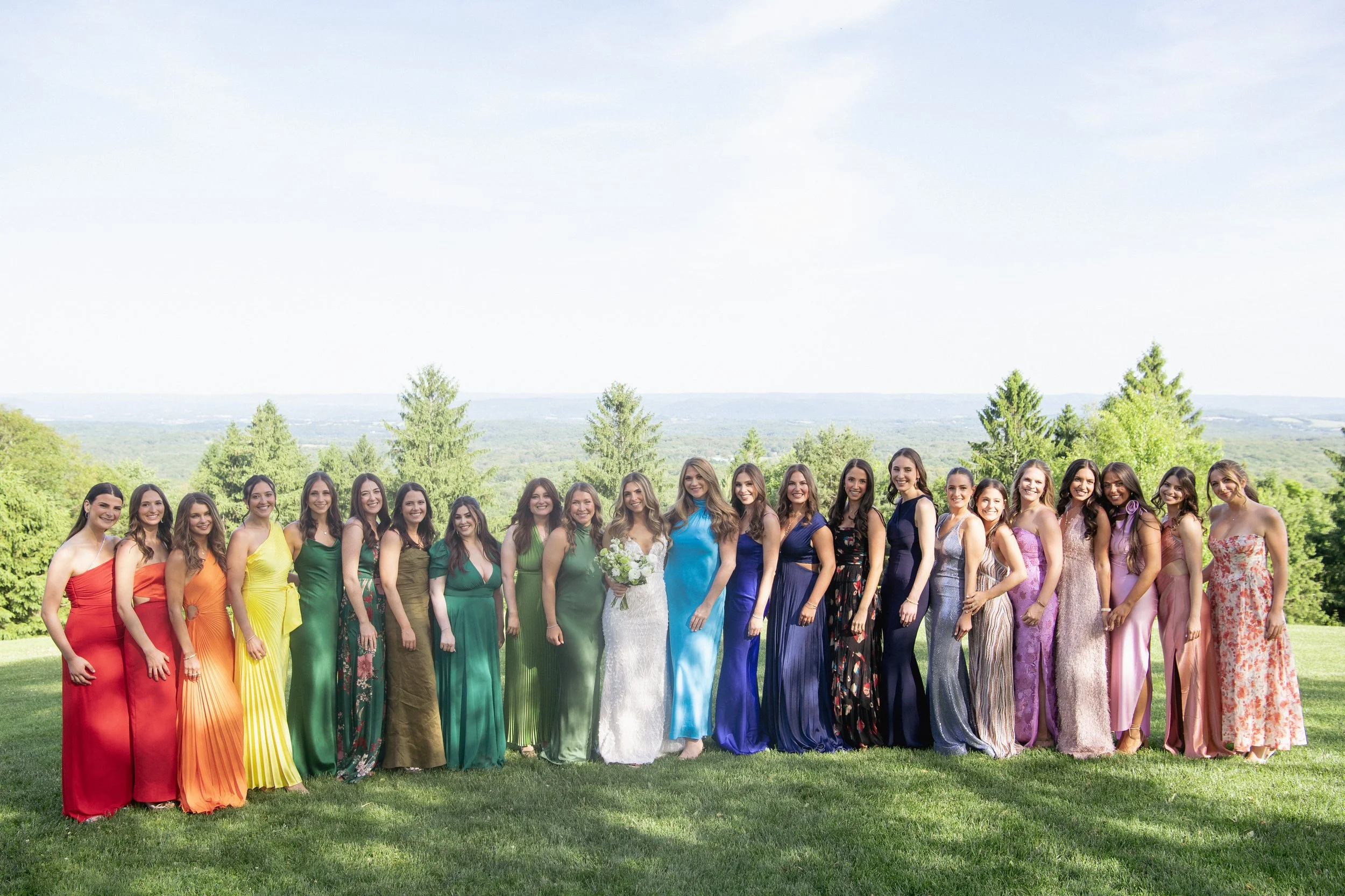 Group of women dressed in colorful gowns standing outdoors on grass with trees and hills in the background, likely at a wedding or special event.