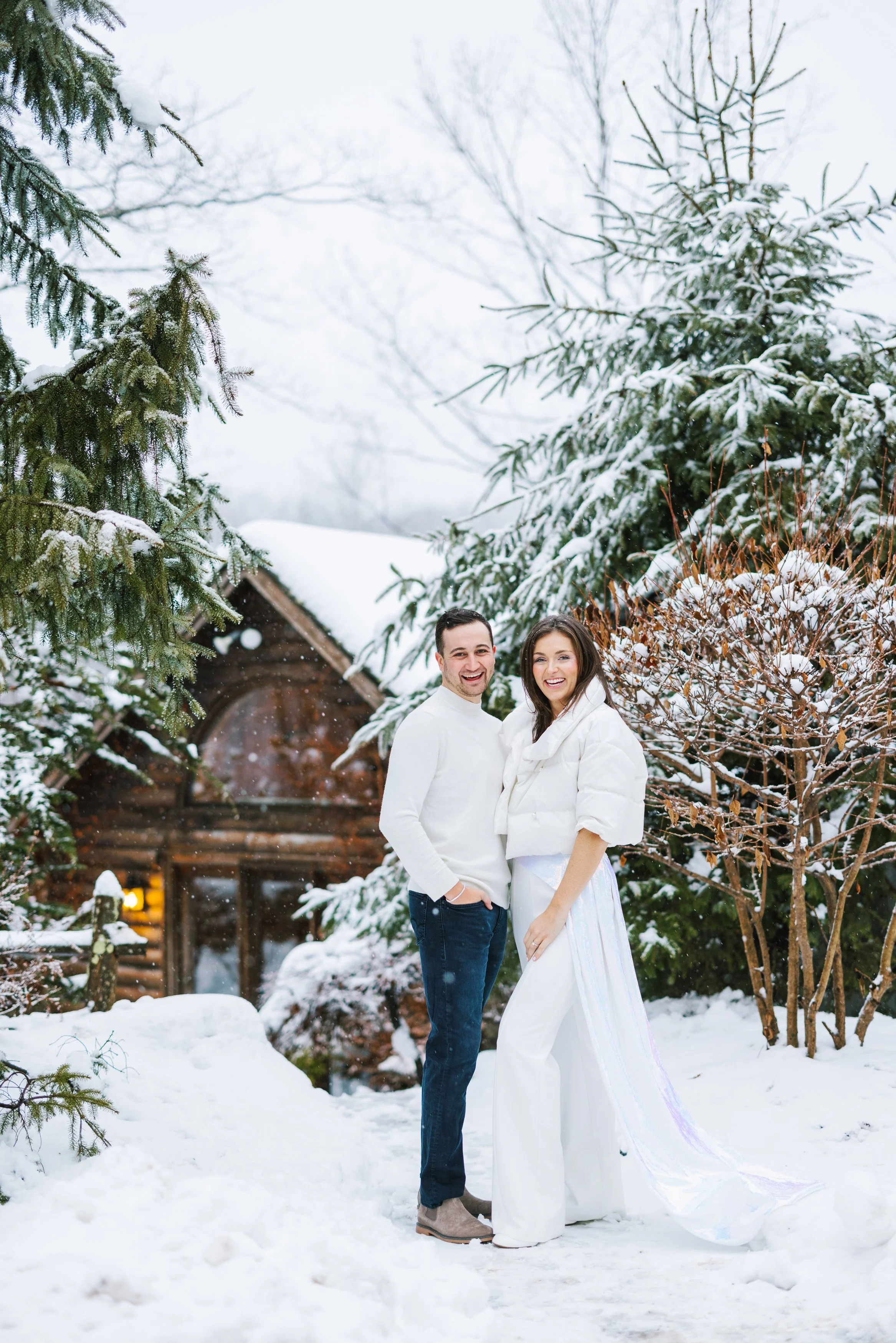 A happy couple dressed in white winter clothing standing in snowy outdoor setting with snow-covered trees and a log cabin in the background.