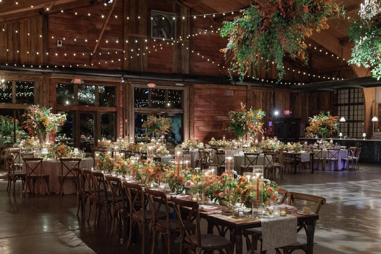 A decorated banquet hall with long tables, floral centerpieces, candles, and string lights, wooden walls, and high ceilings, set for a celebration or event.