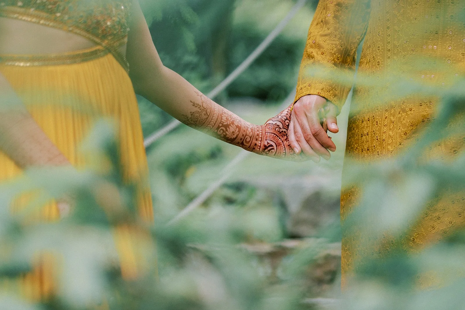 Close-up of a couple holding hands, with the woman wearing traditional henna tattoos on her arm and the man dressed in a yellow embroidered outfit, surrounded by greenery.
