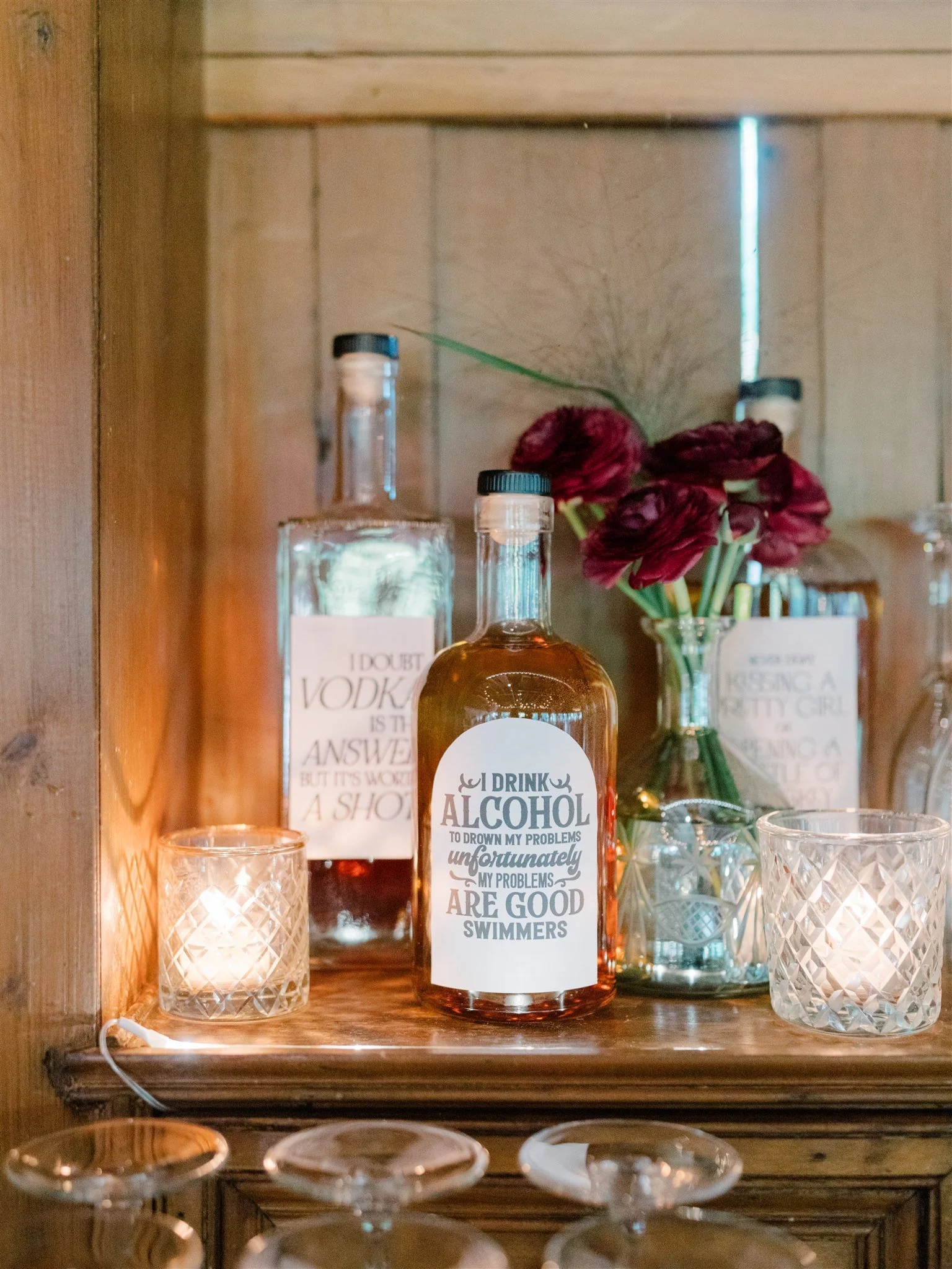 A wooden shelf with two lit candles, a bottle of alcohol with a humorous label, and a vase of dark red flowers.