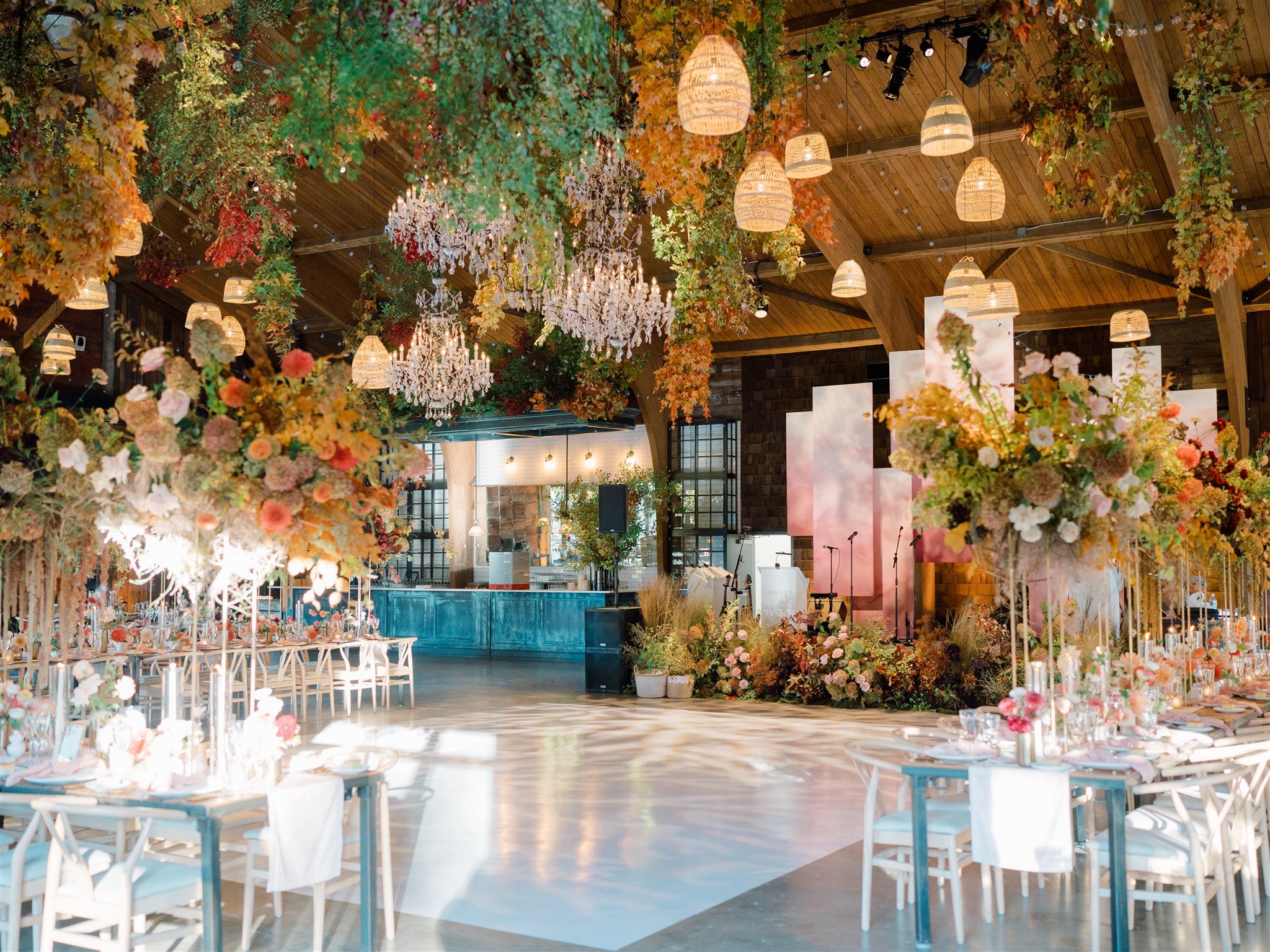 Elegant wedding reception hall decorated with abundant floral arrangements, hanging chandeliers, and warm lighting, set up with tables and chairs for guests.