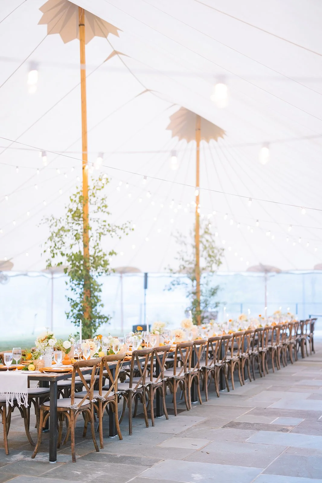 Long outdoor table set for a celebration under a large white tent with wooden poles, decorated with flowers, candles, and string lights, on a stone patio.