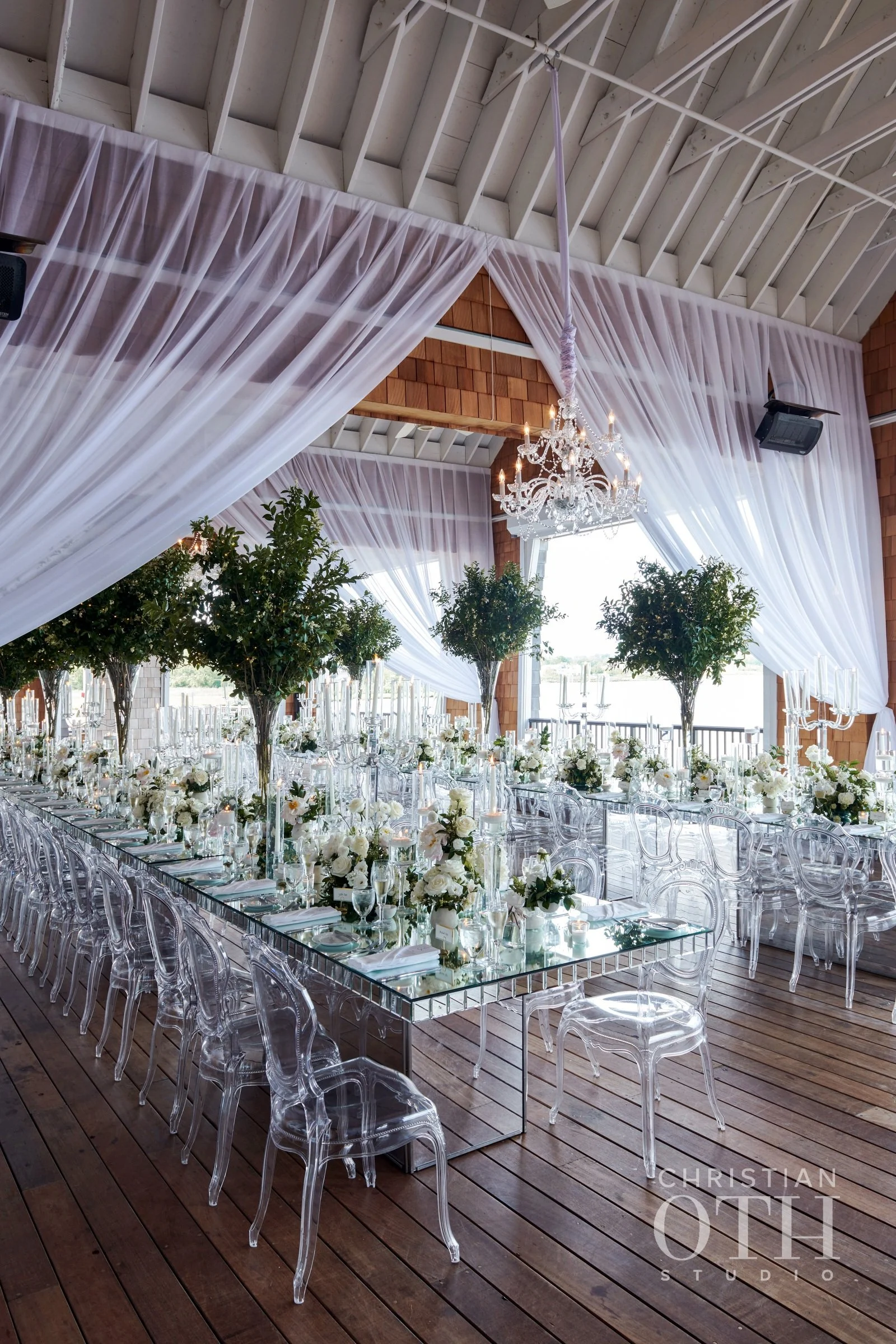 Elegant wedding reception setup with long tables covered in white floral arrangements, surrounded by clear acrylic chairs, under a high ceiling with draped white curtains and a chandelier.