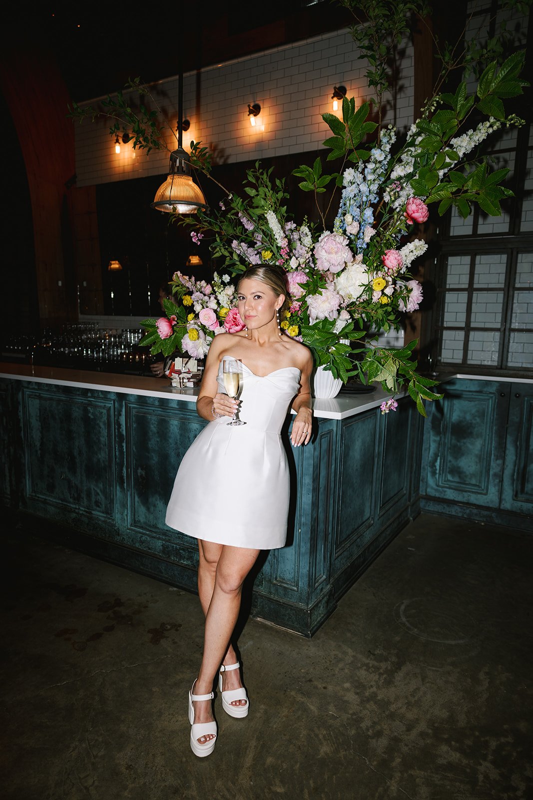 A woman in a white strapless dress with a champagne glass standing next to a large floral arrangement with pink and white flowers and green leaves, in a dimly lit indoor space with warm lighting and a dark blue bar counter.