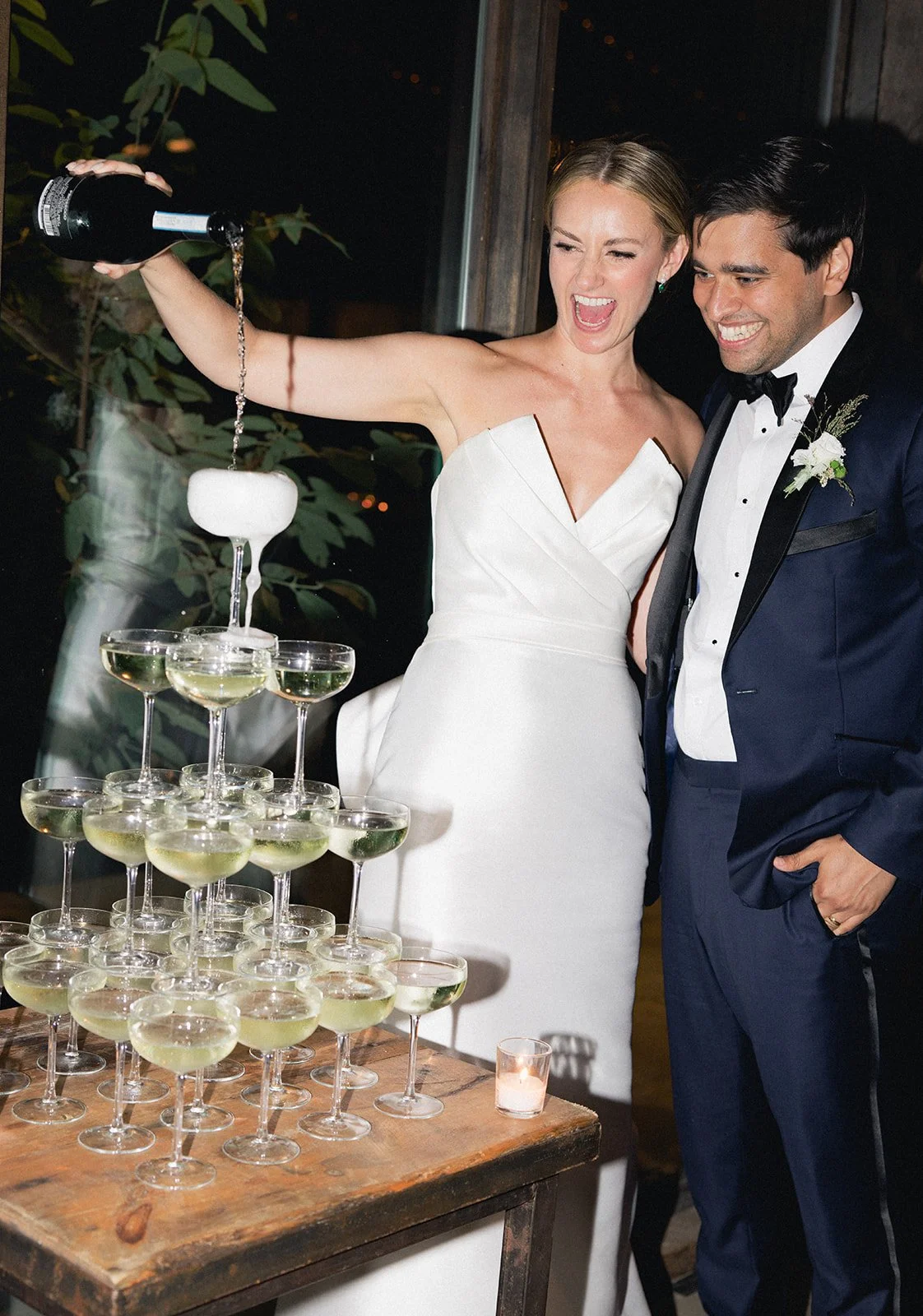 A bride and groom celebrating at their wedding, with the bride pouring champagne into a tower of glasses, both smiling and dressed in formal wedding attire, indoors at night.