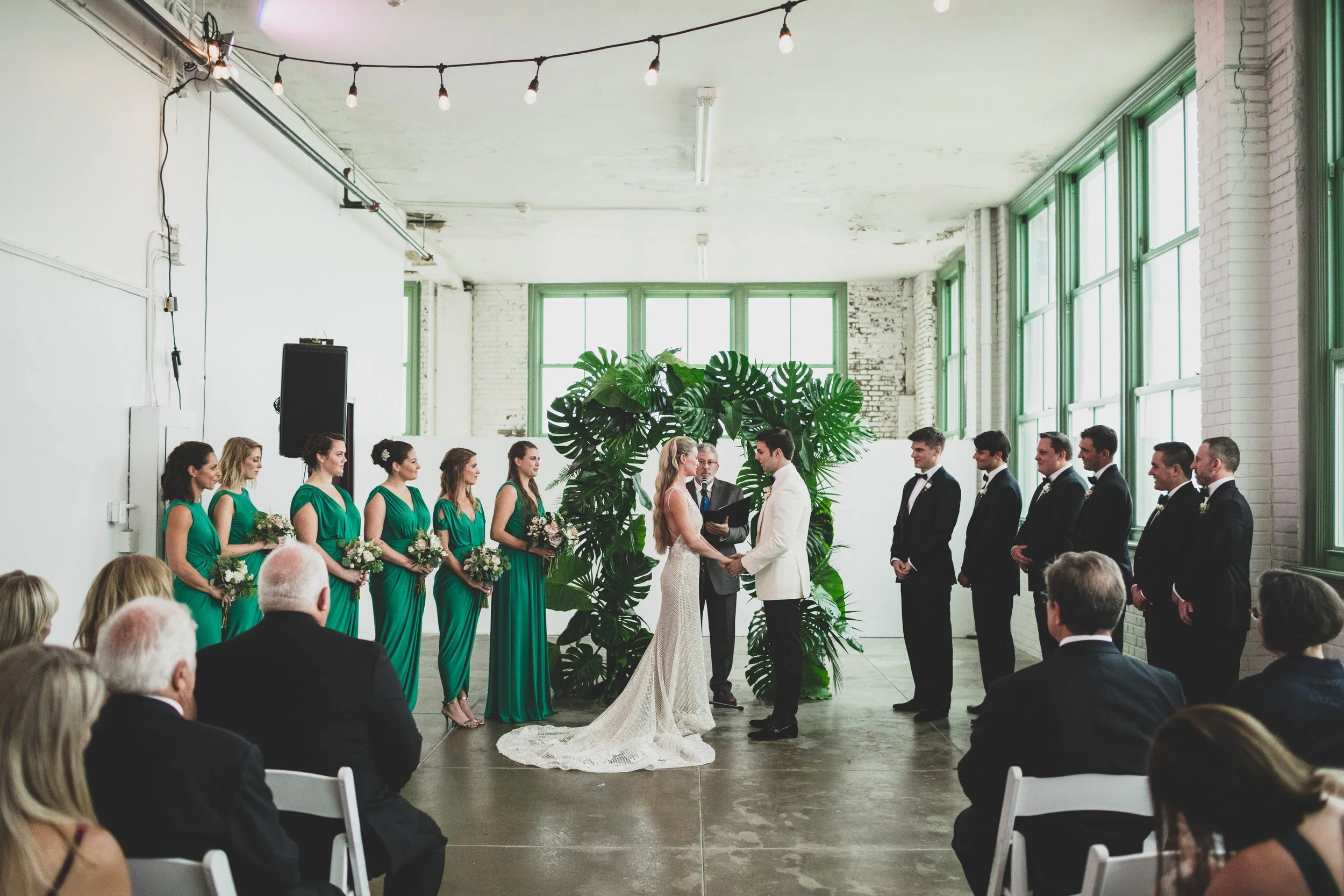 A wedding ceremony taking place indoors with the bride and groom holding hands in front of an officiant, flanked by bridesmaids in green dresses on one side and groomsmen in black tuxedos on the other, with guests seated and watching.