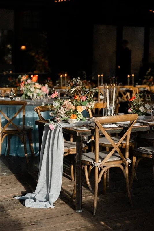 A banquet table decorated with floral centerpieces, candles, and a draped tablecloth in a dimly lit event space.