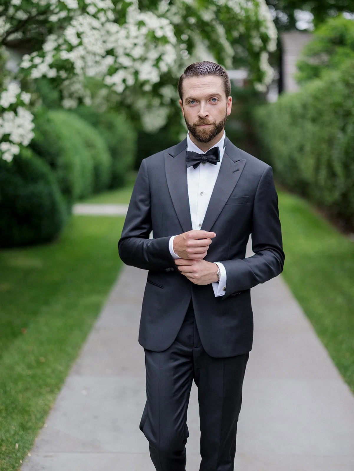 A man in a black tuxedo with a bow tie standing on a sidewalk in a garden, looking at the camera.