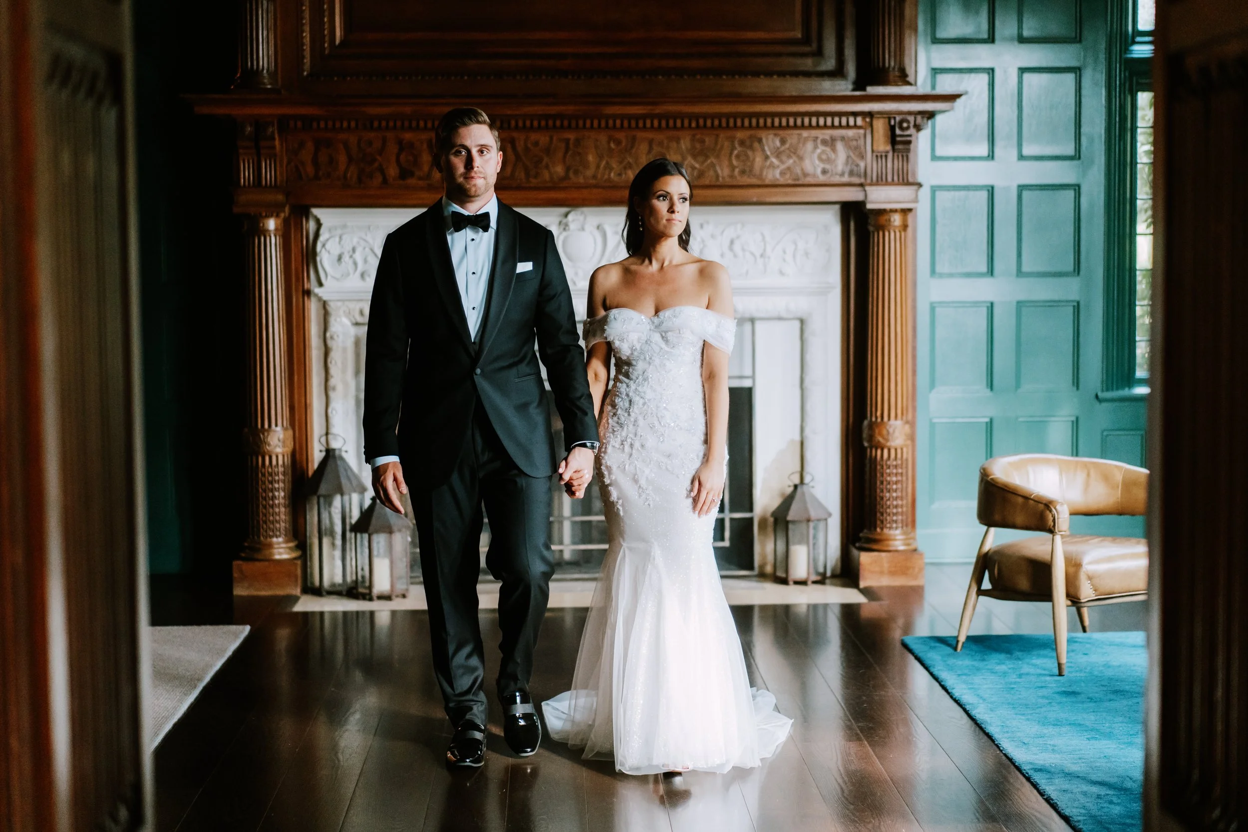 A bride and groom holding hands indoors with wooden decor and a fireplace in the background. The bride is in a white off-shoulder wedding gown, and the groom is in a black tuxedo with a bow tie.