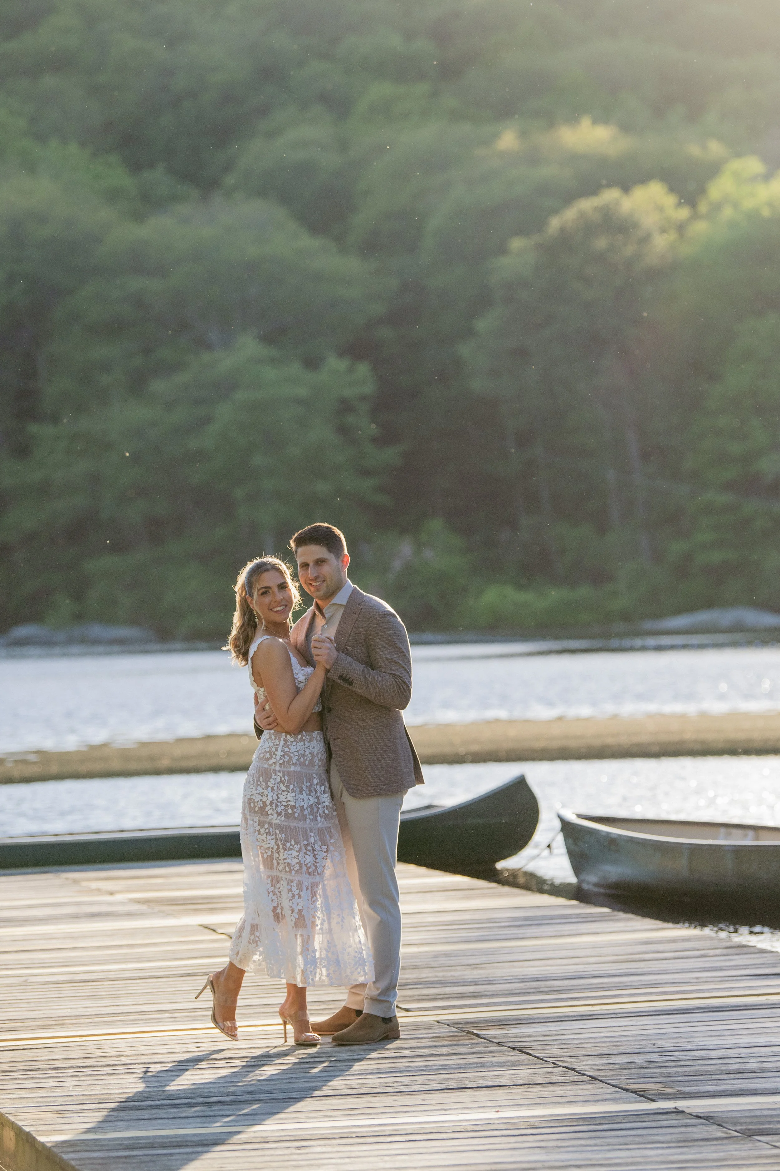 A couple in wedding attire dancing on a wooden dock near a lake, with canoes and a forested shoreline in the background during sunset.