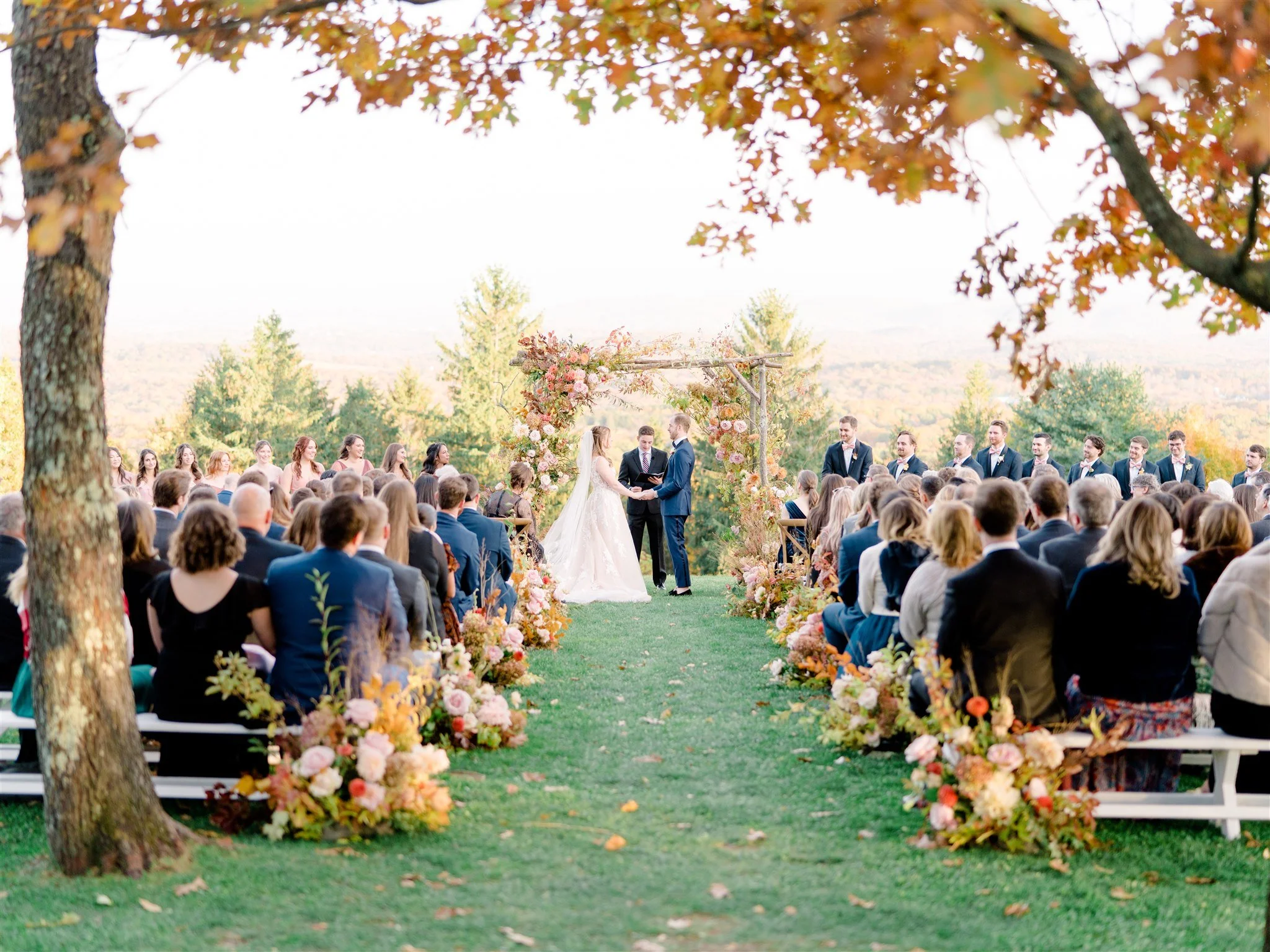 Outdoor wedding ceremony with the bride and groom exchanging vows under a floral arch, surrounded by guests seated on wooden benches, with trees and a scenic view in the background.