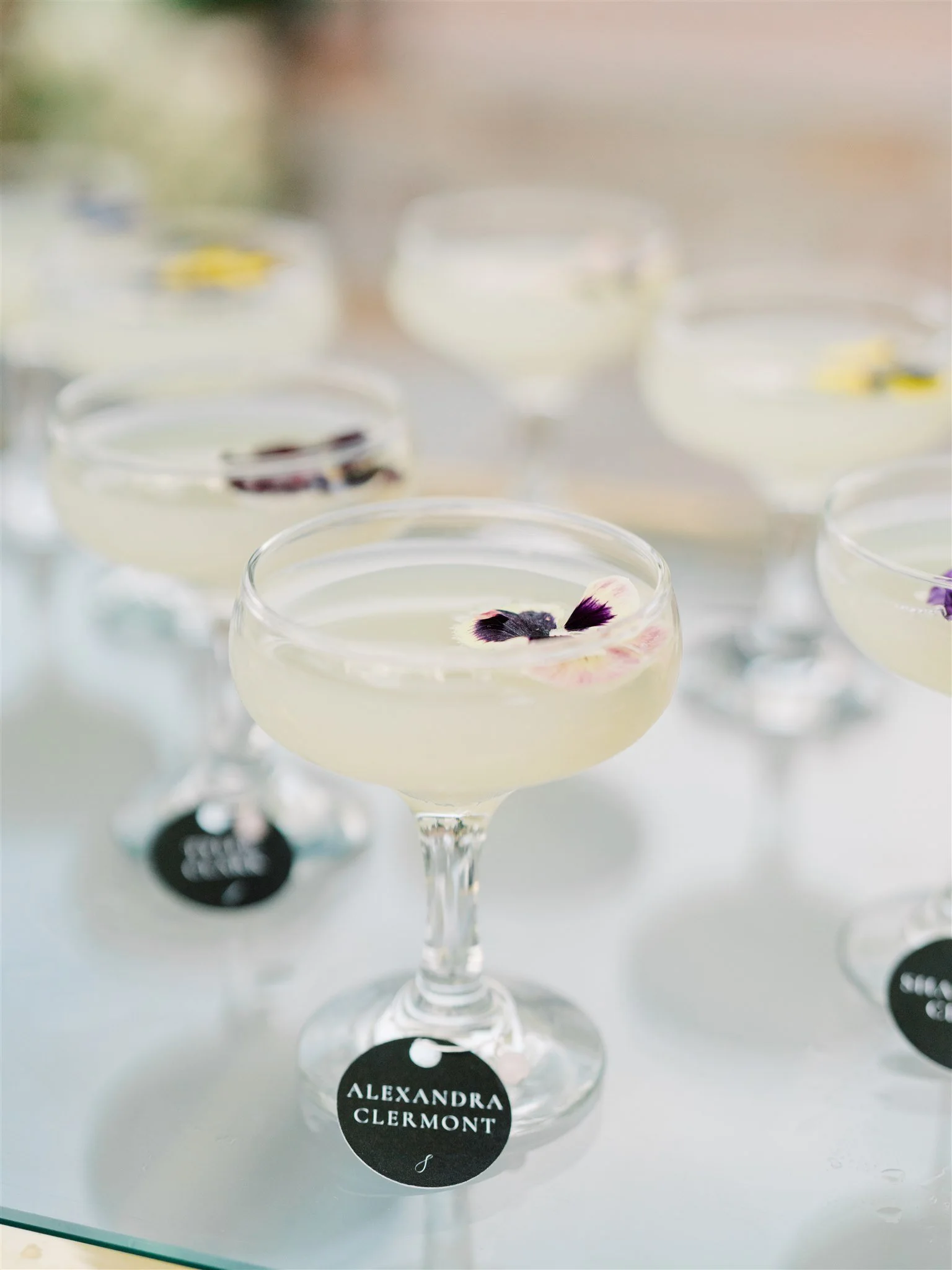 Elegantly arranged cocktail glasses with pale yellow drinks garnished with purple and white flower petals, on a white surface.