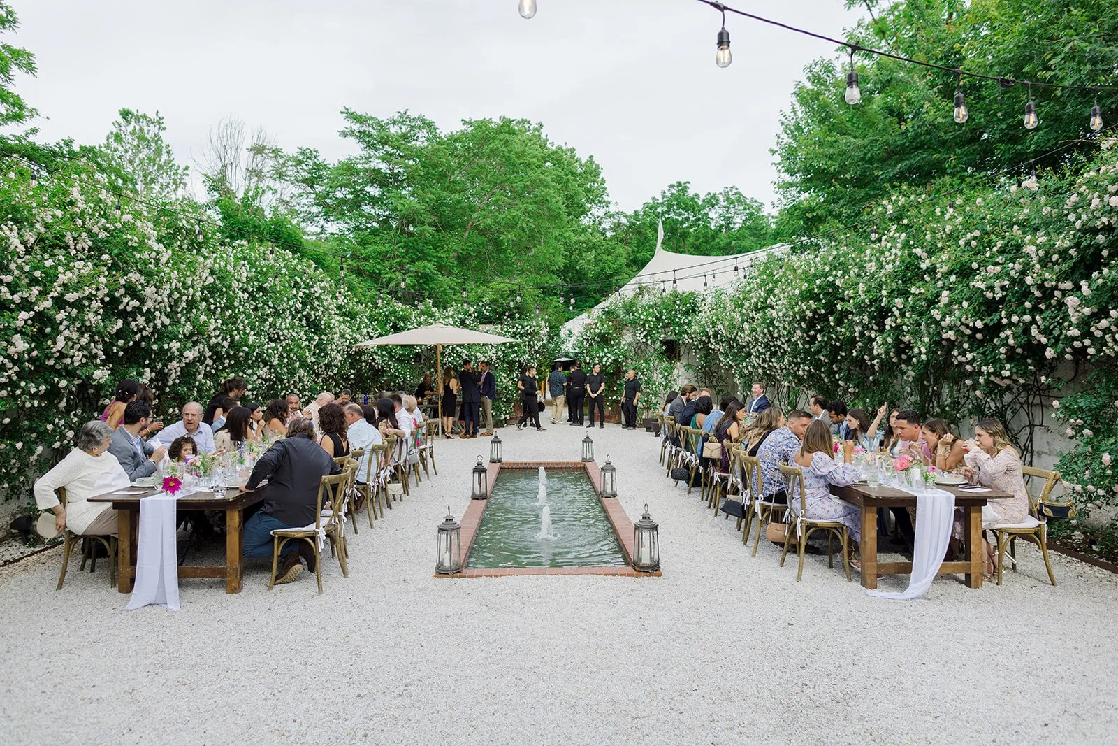 Outdoor wedding reception with long tables arranged on a gravel pathway, surrounded by lush green trees and flowering bushes, with string lights overhead and a central water feature bordered by lanterns.