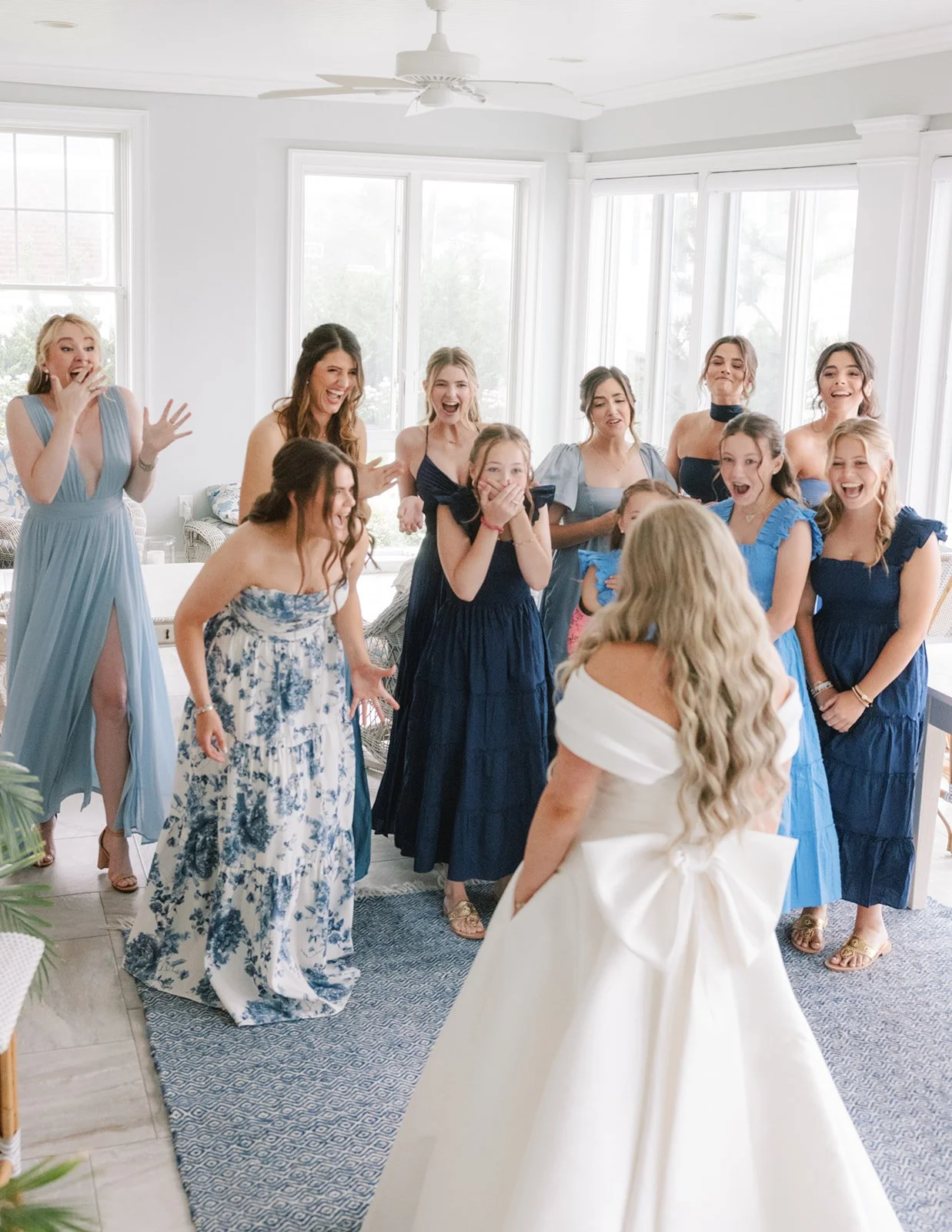 A group of women in a bright living room, some in blue dresses, surprise and cheer as they look at a woman in a white wedding dress facing away from the camera.