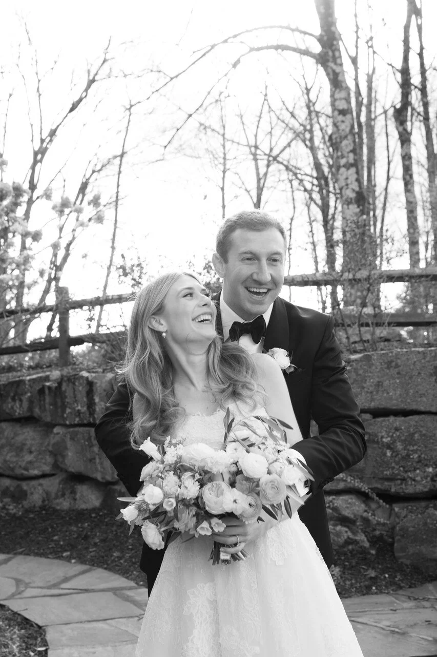 Black and white photo of a bride and groom smiling and embracing outdoors, with the bride holding a bouquet of flowers.