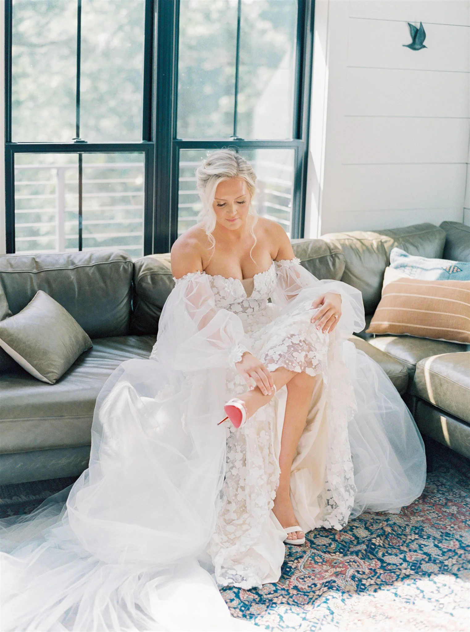 A bride in a wedding dress sitting on a sofa in a bright room, putting on a white high heel shoe.