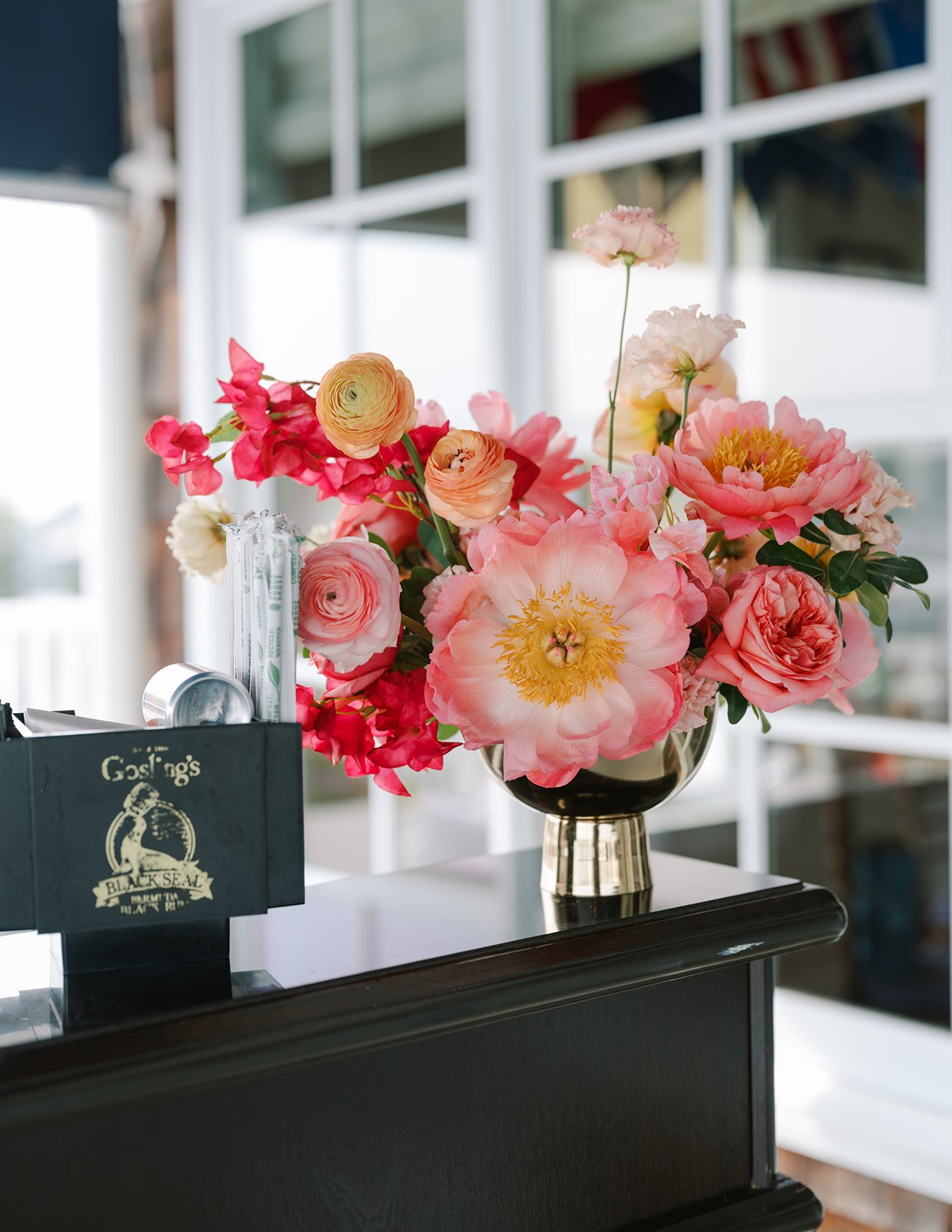 A large bouquet of pink, white, and peach flowers in a black and gold vase on a black counter, with a black box and some miscellaneous items nearby, in front of a window with a grid pattern.