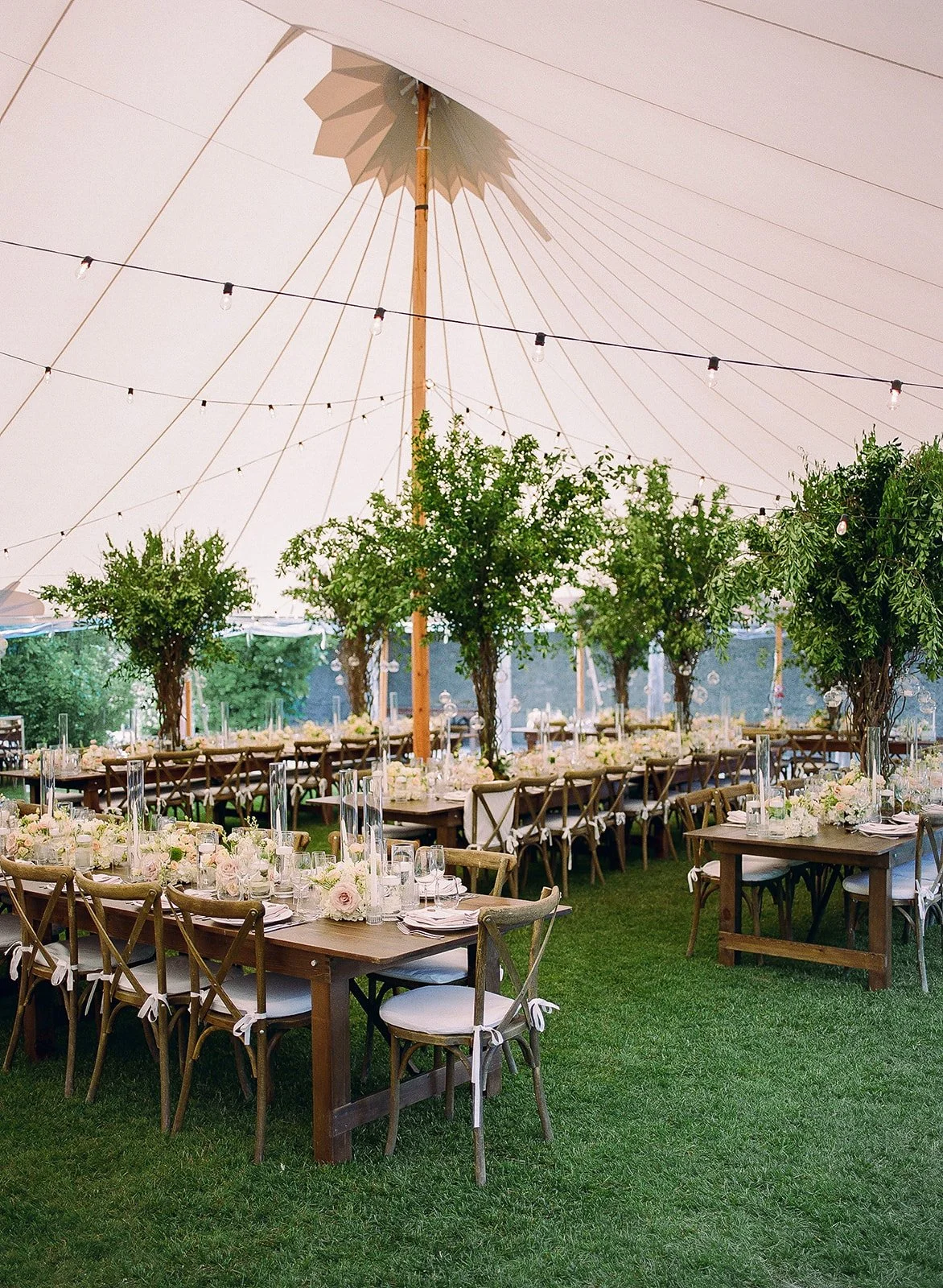 Outdoor wedding reception setup under a large white tent with wooden tables, chairs, floral centerpieces, string lights, and trees.