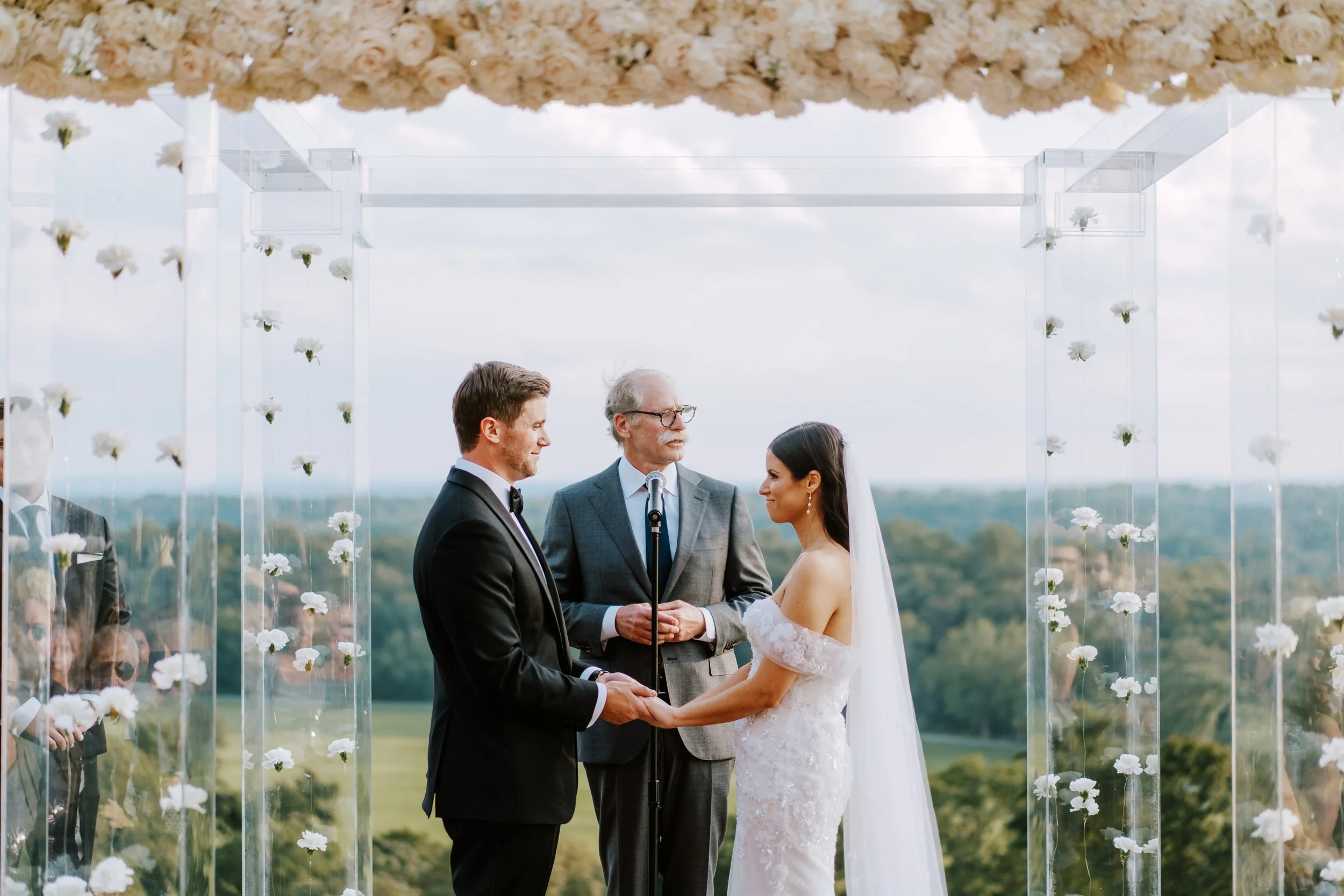 A bride and groom exchanging vows at outdoor wedding ceremony with officiant, holding hands and looking at each other, with a scenic landscape in the background.