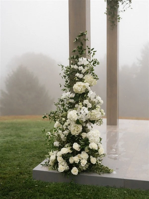 A floral arrangement with white roses, hydrangeas, and greenery on a platform outside, with foggy background and wooden posts.